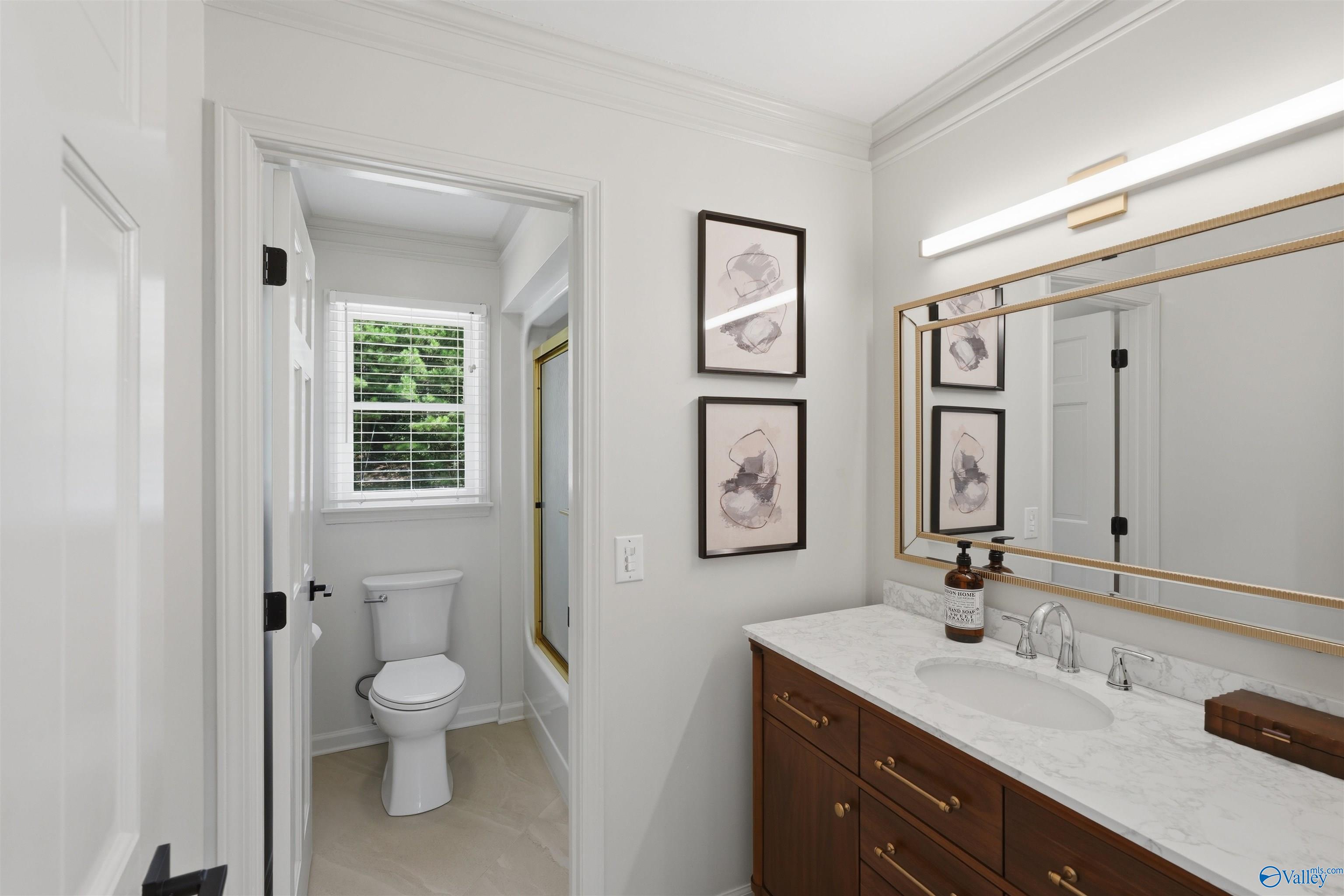 Bathroom vanity with marble-look counter and mirrors beside a separate toilet area with a window.