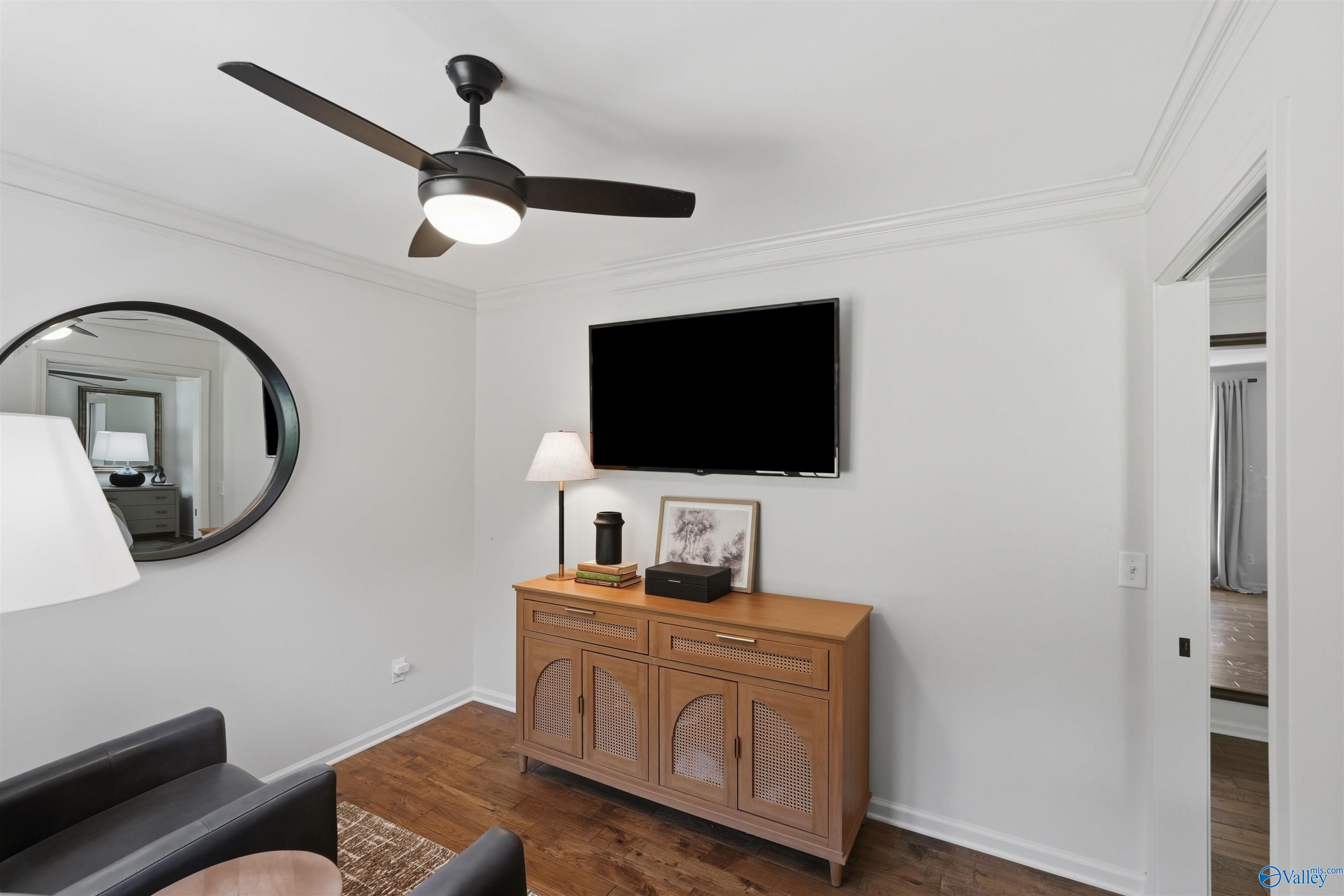 Furnished room with a wall-mounted television, wooden cabinet, round mirror, and black ceiling fan.