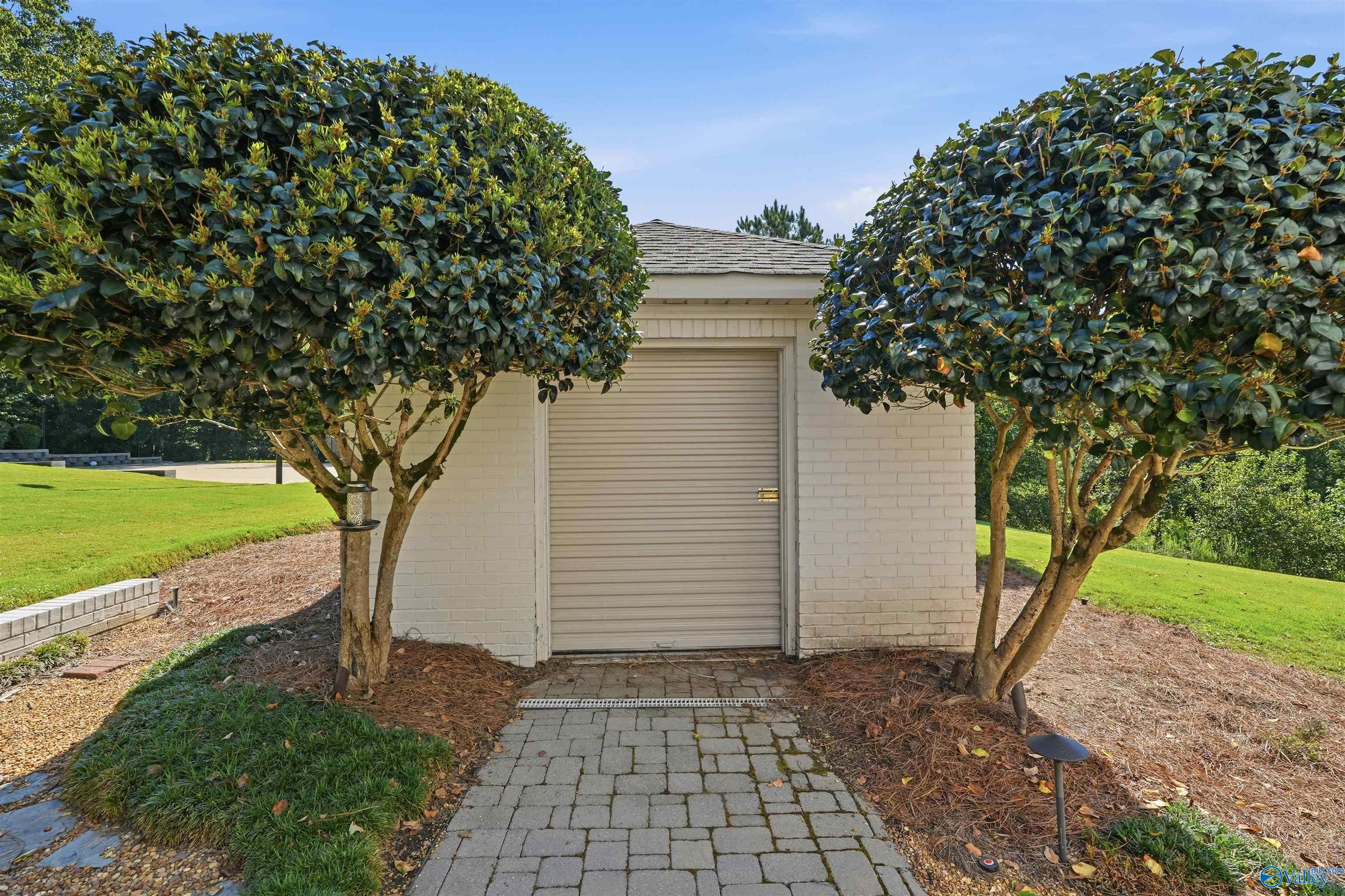 Light-brick outbuilding with a closed roll-up garage door between two trimmed trees.