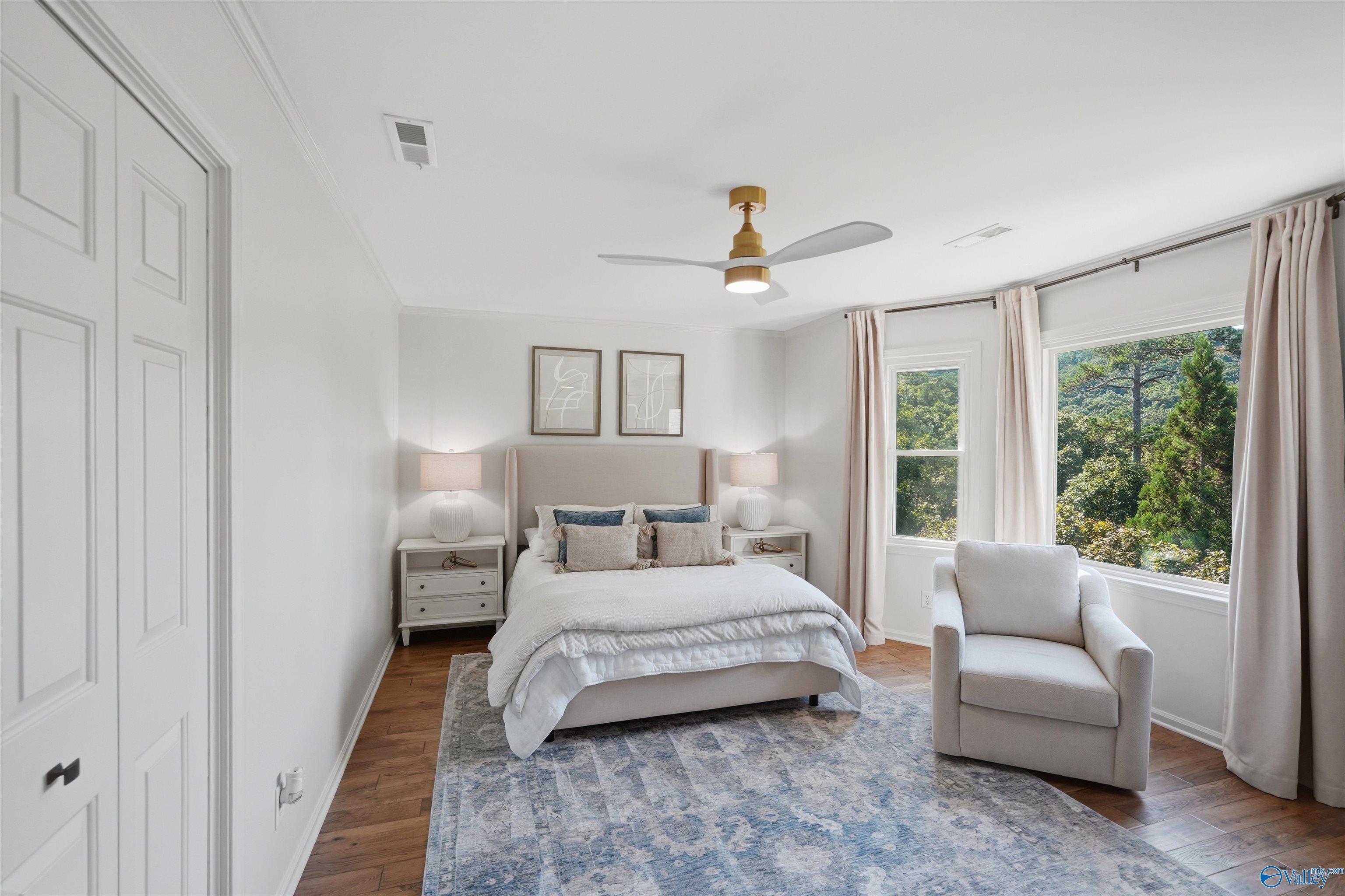Staged bedroom with an upholstered bed, blue patterned rug, and armchair beside large windows.
