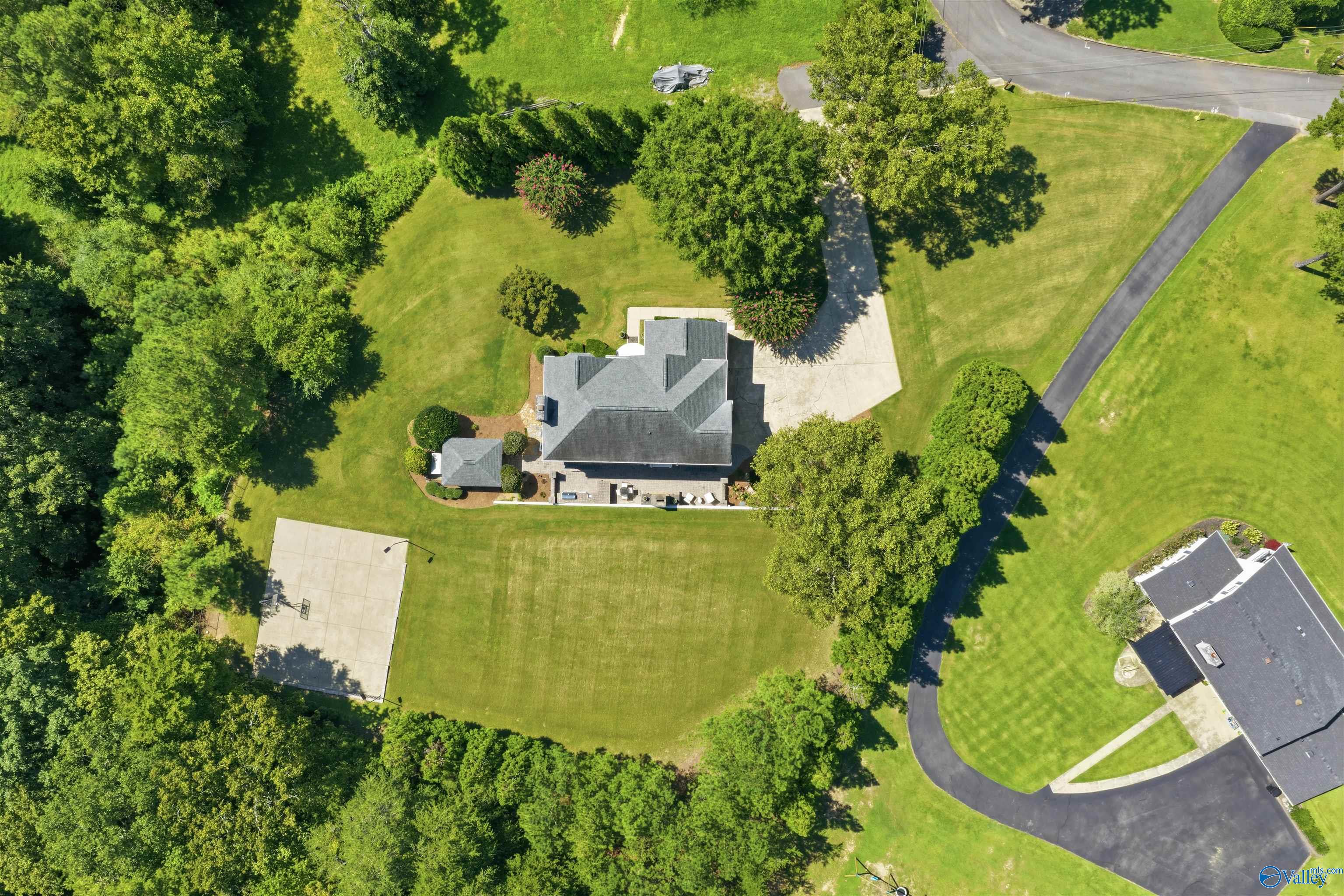Aerial view of a gray-roofed home with a large lawn, mature trees, and a basketball court.
