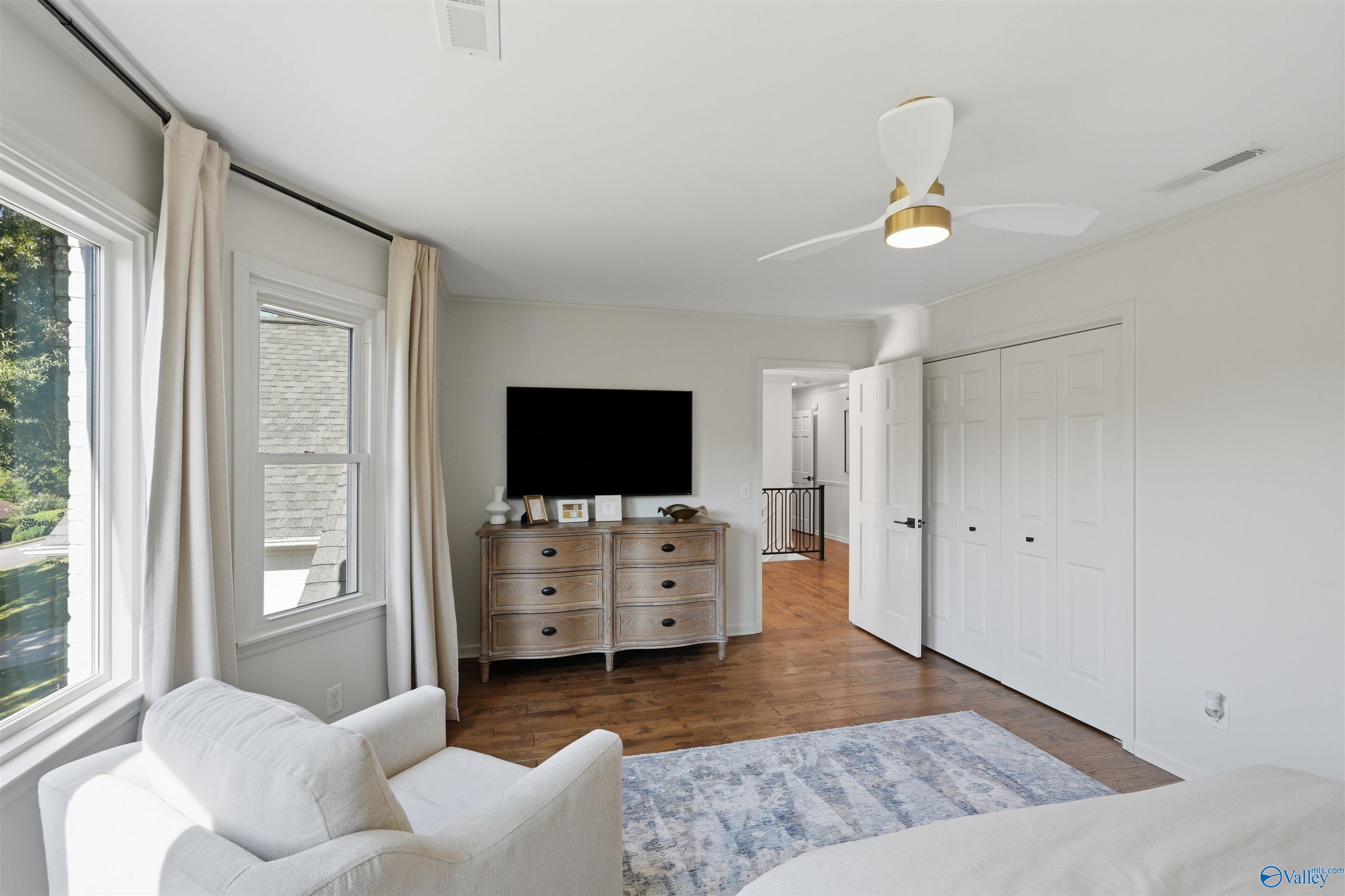 Bedroom with a wooden dresser and TV, white paneled closet doors, and large curtained windows.