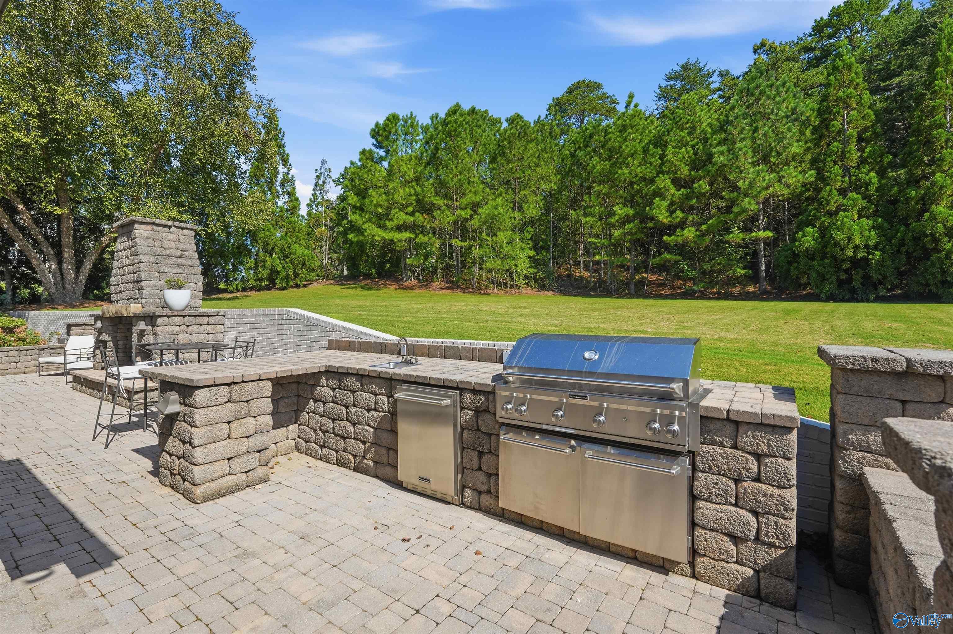 Stone outdoor kitchen with stainless steel grill and sink on a paved patio beside a tree-lined lawn.