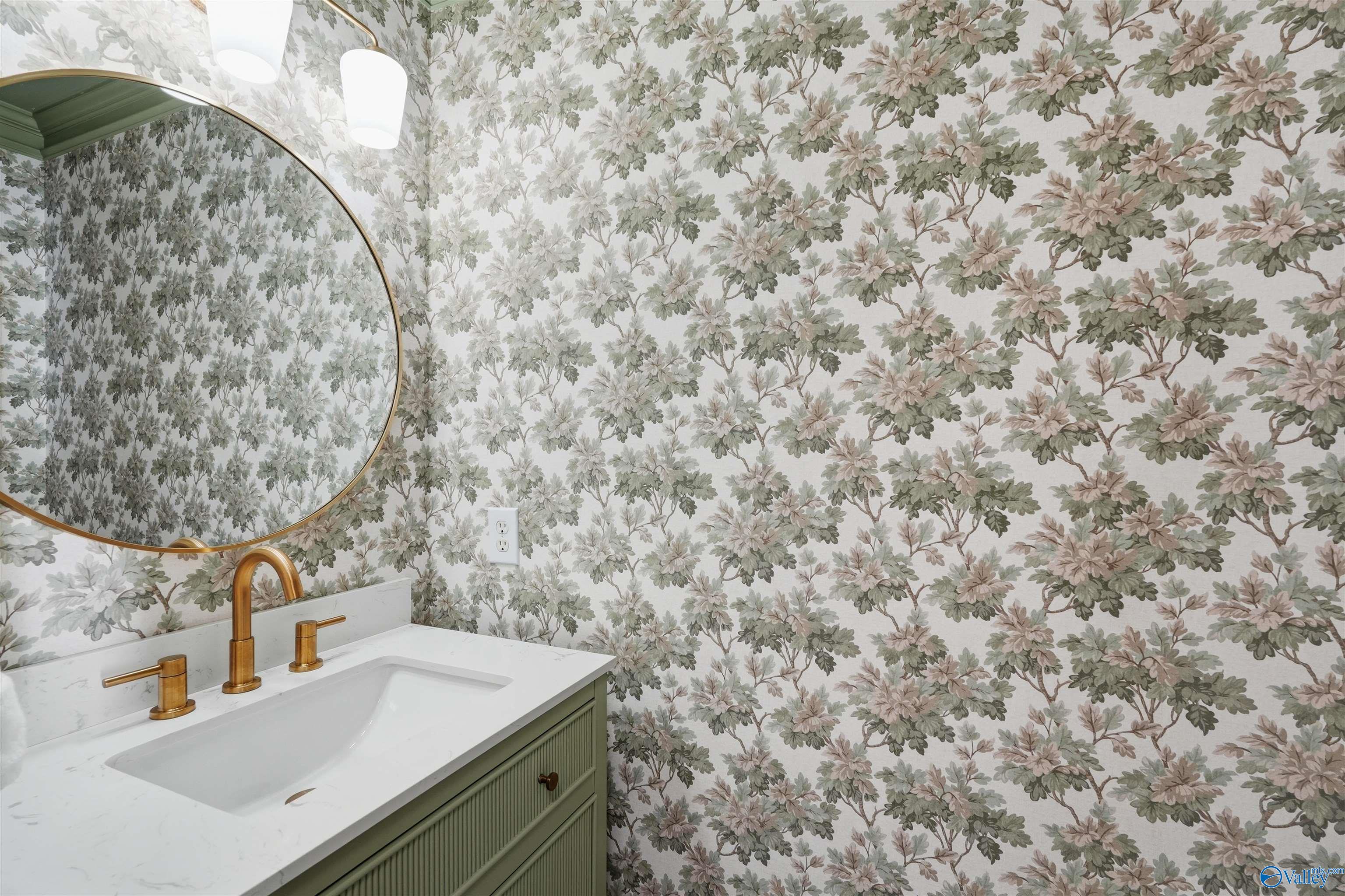 Bathroom vanity with a brass faucet, round mirror, sage green cabinet, and floral wallpaper.