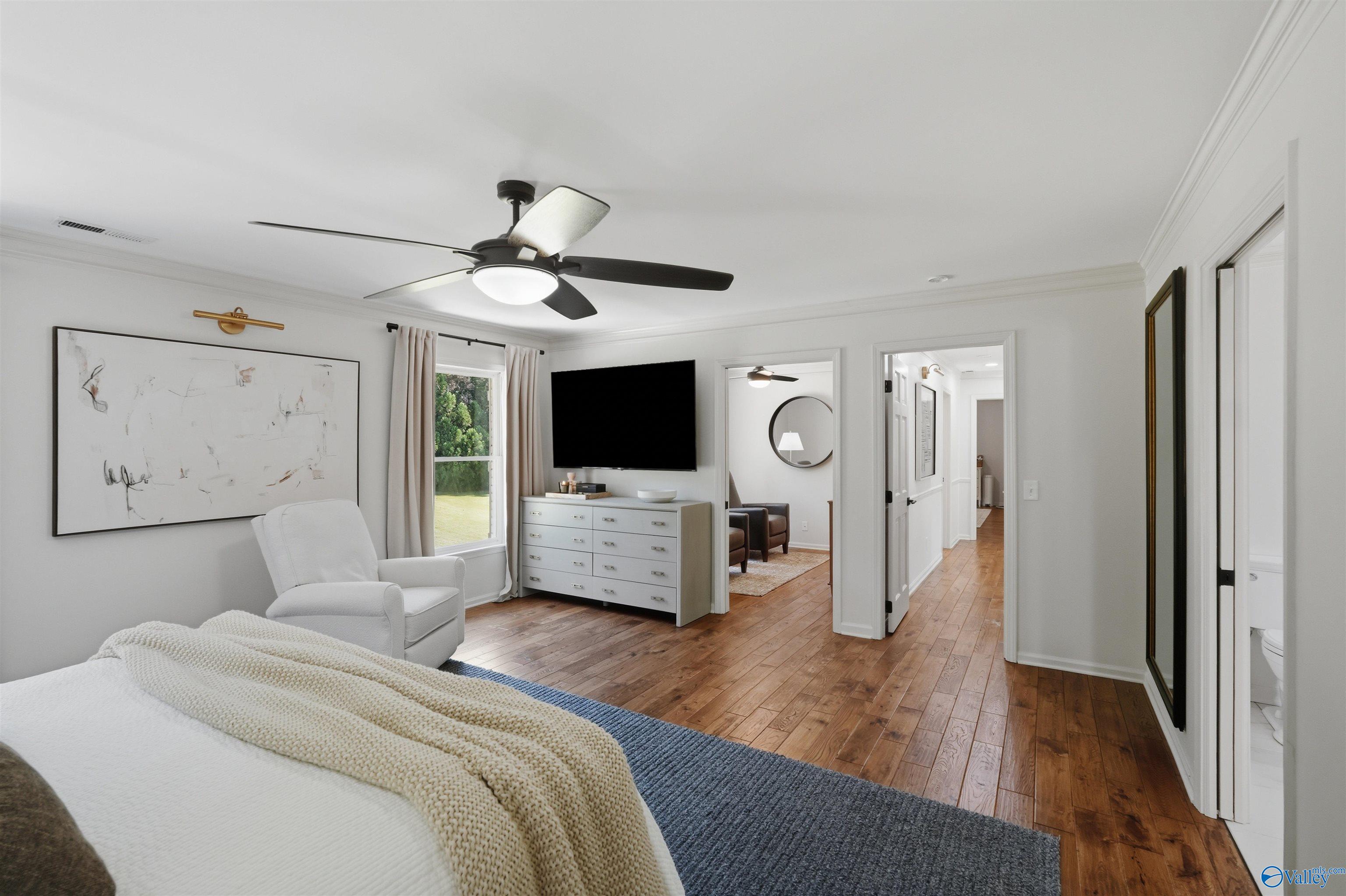 Furnished bedroom with warm wood floors, a white armchair, and a television above the dresser.
