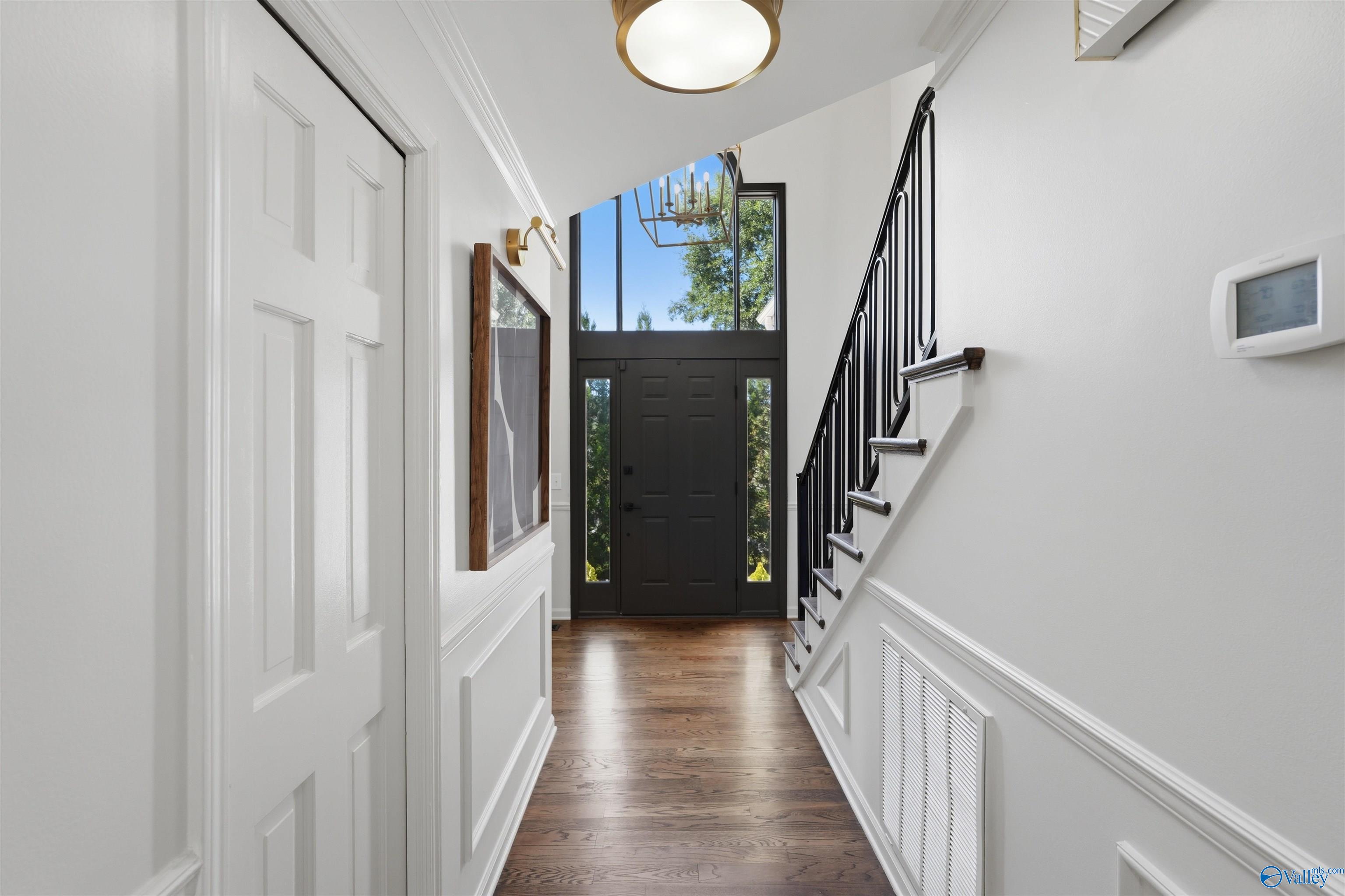 Entry hall with a dark front door, tall windows, hardwood floors, and a staircase with black railing.