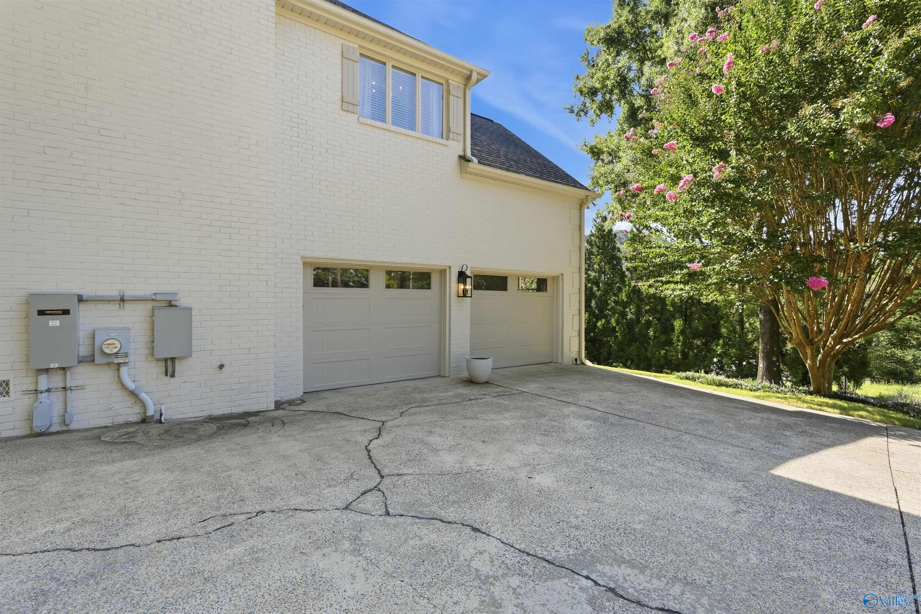 White brick house with two garage doors and a cracked driveway beside a flowering tree.