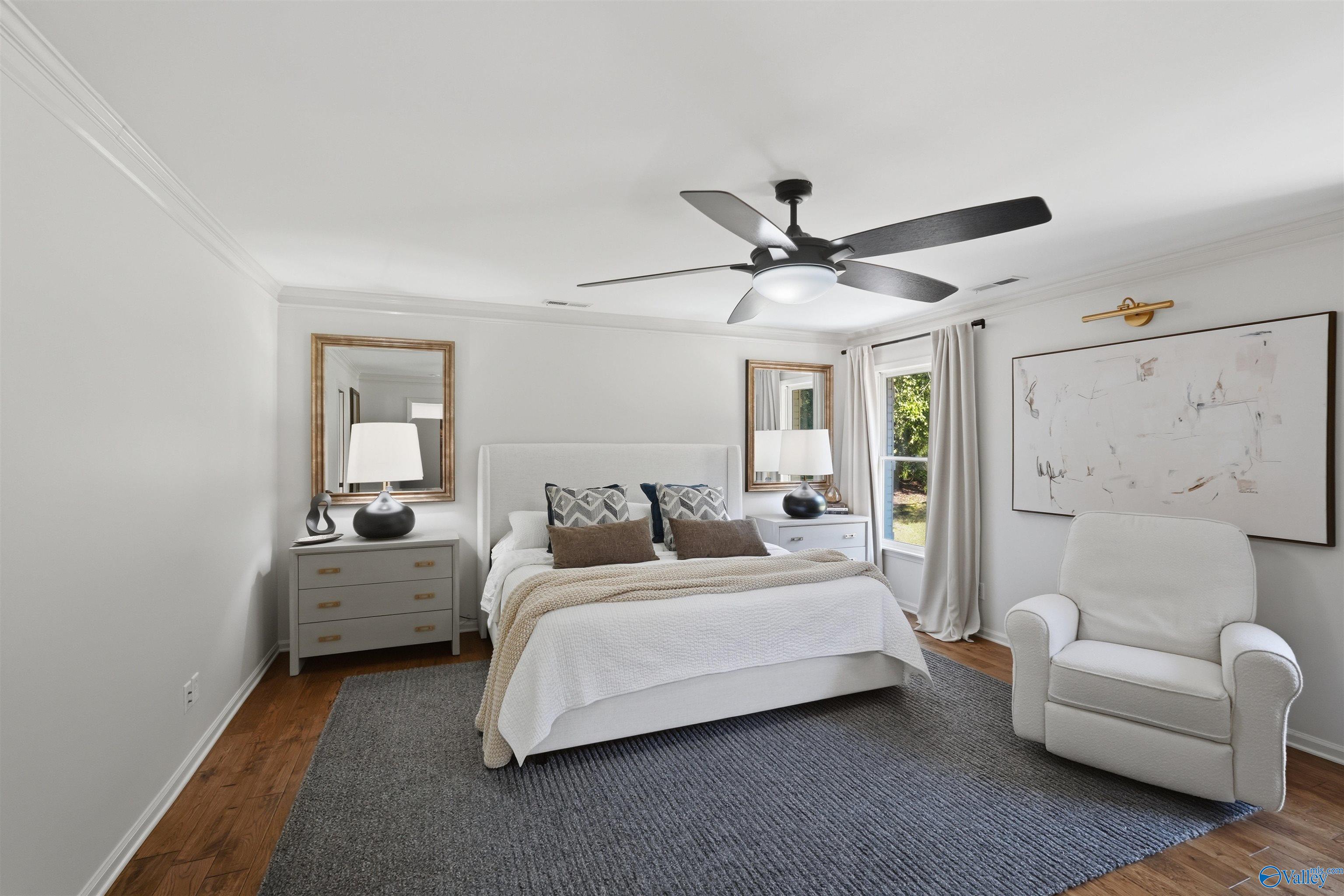 Staged bedroom with a white bed, gray rug, hardwood floors, and a white armchair beside the window.