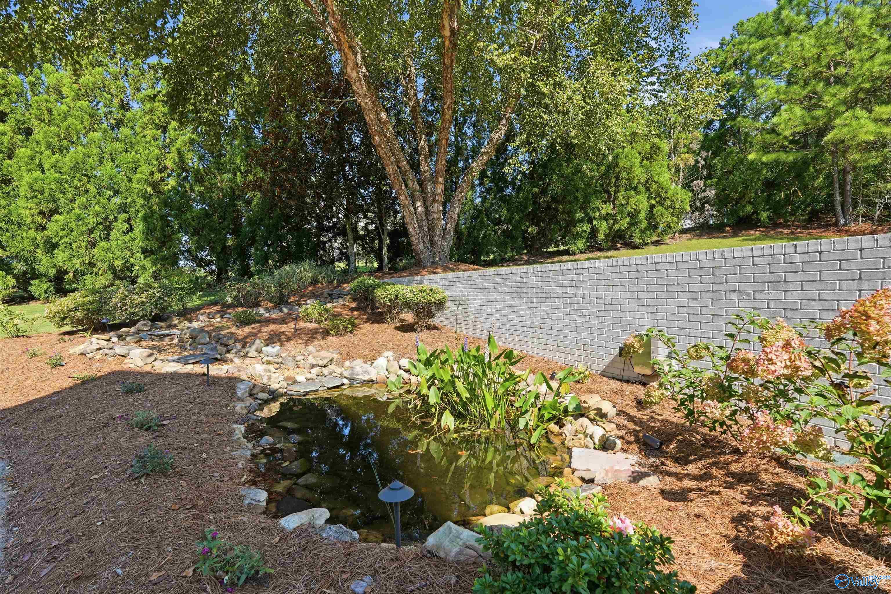 Backyard pond edged with rocks and leafy plants beside a gray brick retaining wall.
