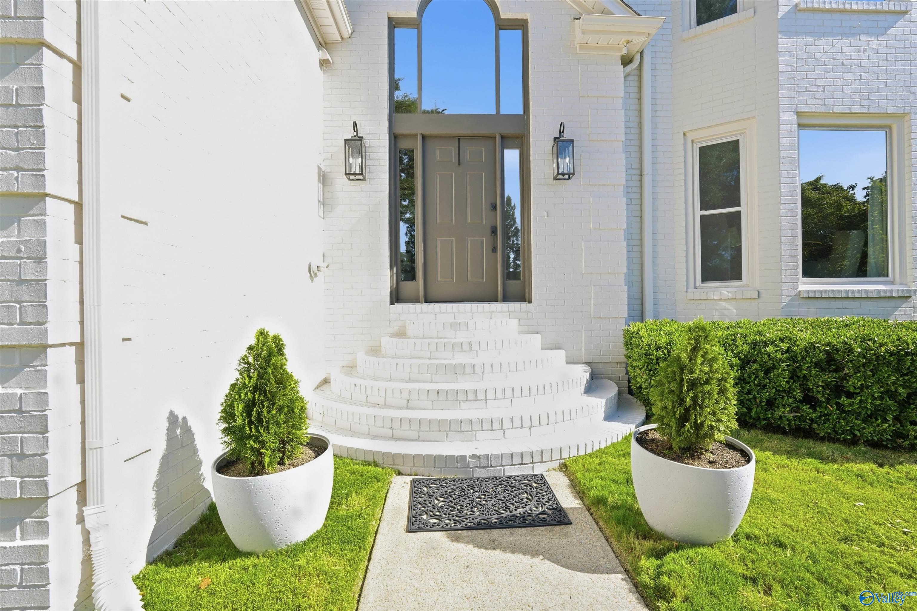 White brick home entrance with curved steps, an arched window, and matching potted shrubs.