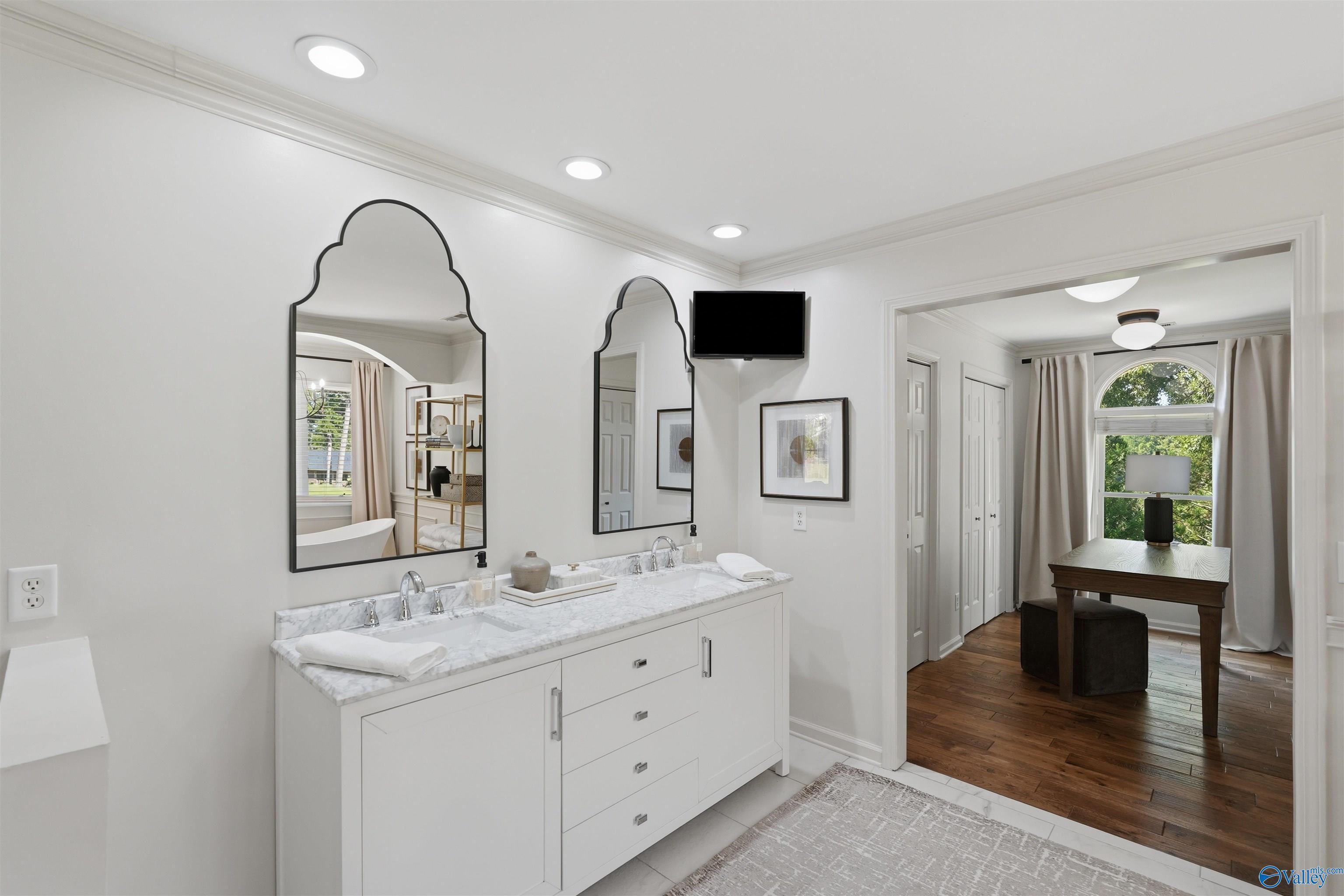 Bathroom with a double-sink marble vanity and arched mirrors, opening to a room with a desk and arched window.