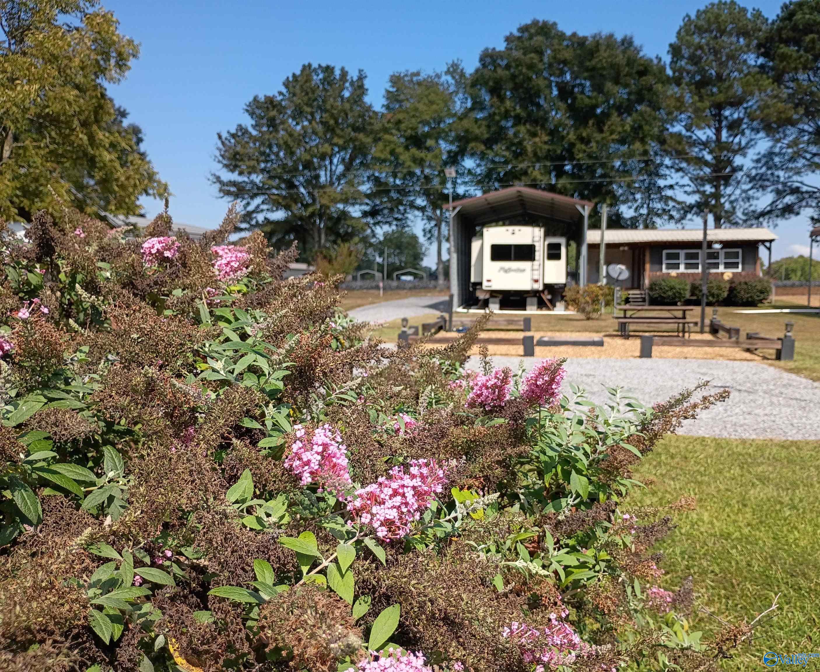 Property exterior with pink flowering shrubs, a gravel drive, an RV carport, and a small building.