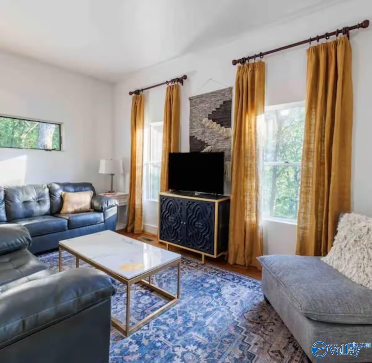 Furnished living room with blue sofas, a patterned rug, and tall windows framed by gold curtains.