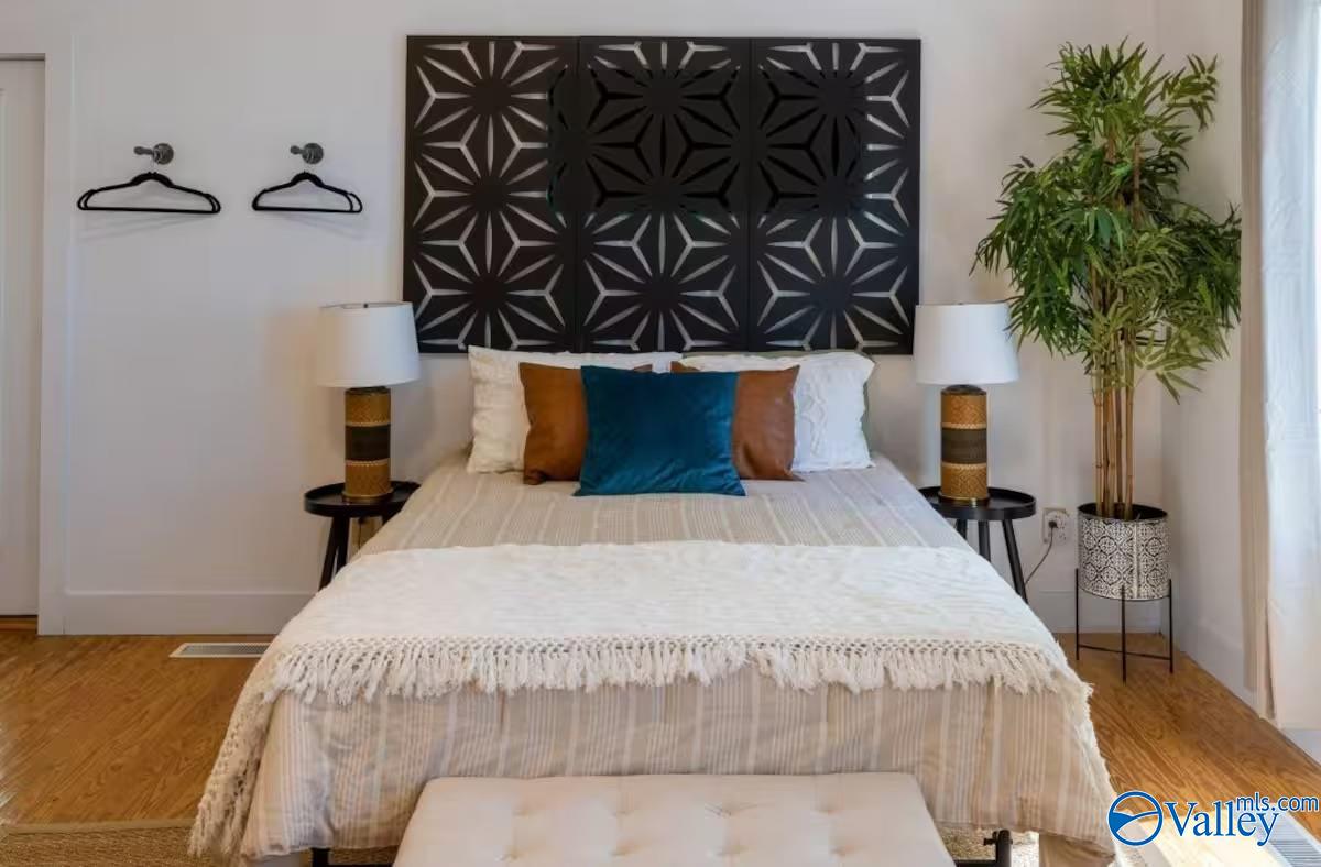 Staged bedroom with a centered bed, geometric wall panel, matching lamps, and a tall potted plant.