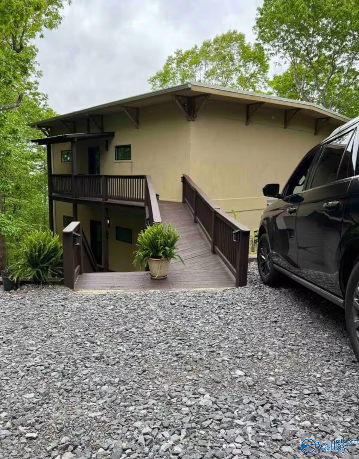 Tan two-level home with a raised wooden entrance ramp, gravel driveway, and surrounding trees.