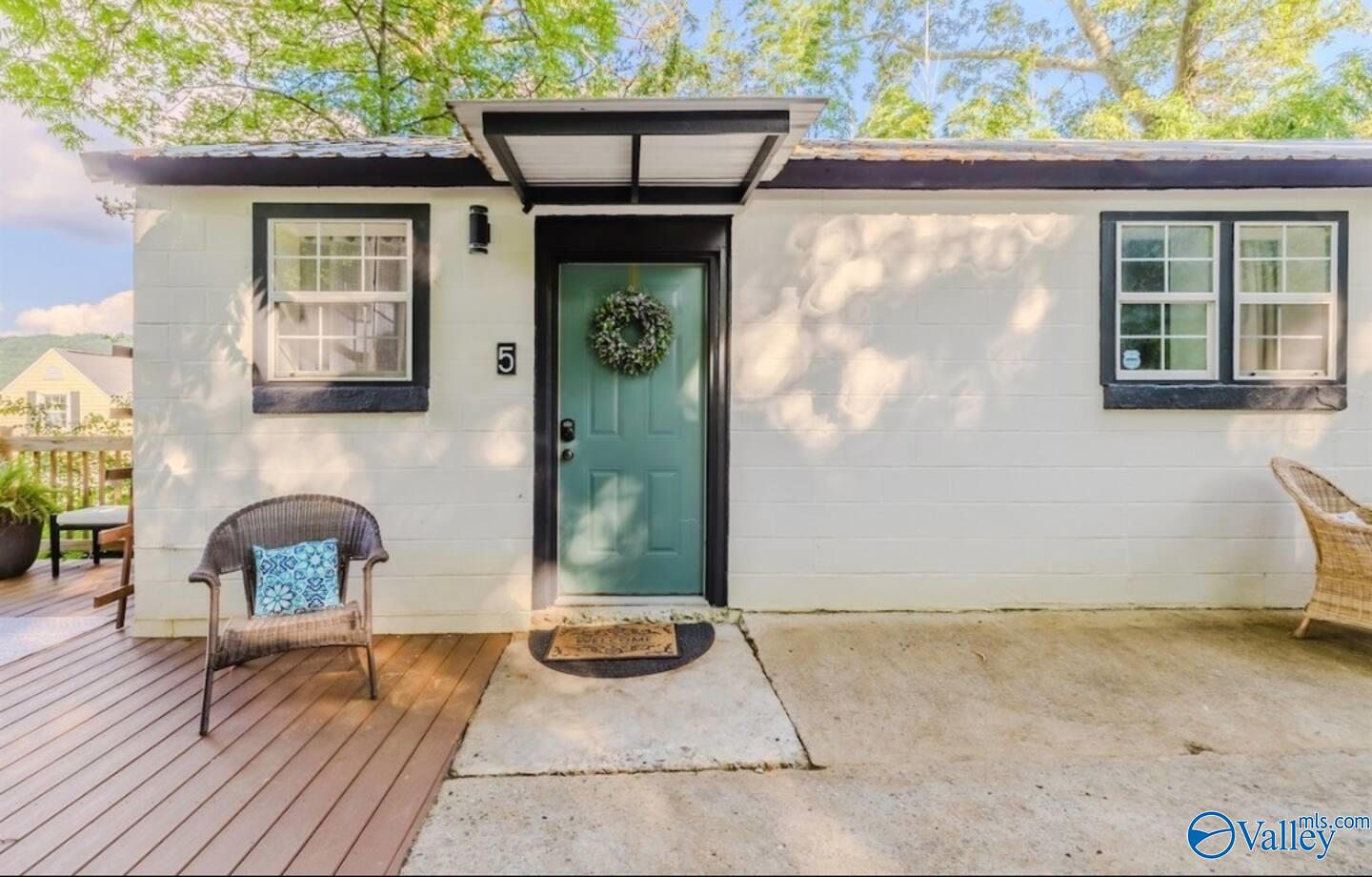 White home entrance with a teal door, wreath, black trim, and wicker chairs on the deck.