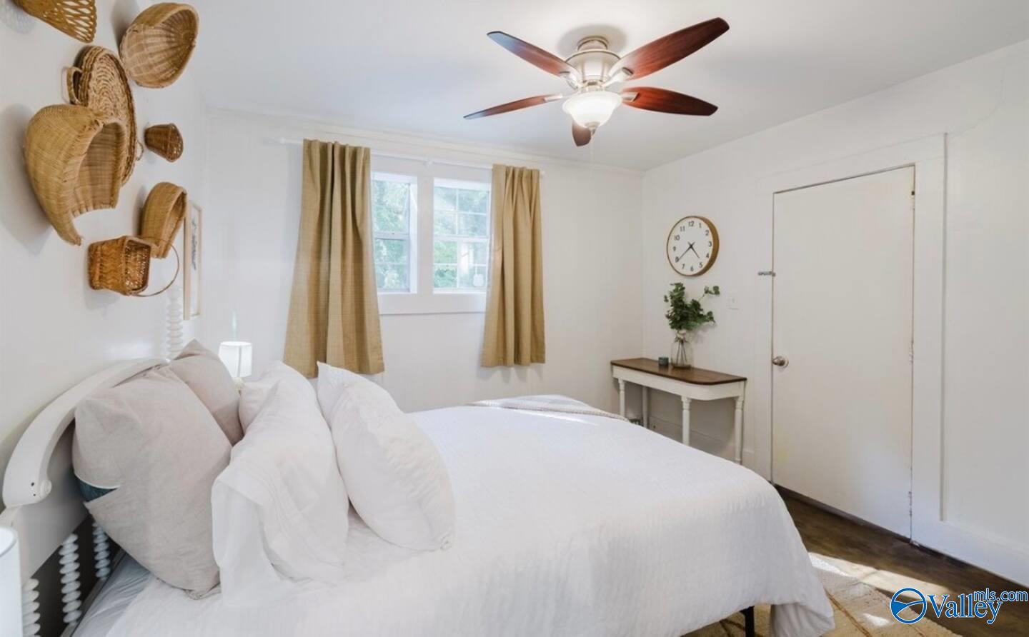 Bedroom with a white bed, woven wall baskets, tan curtains, and a dark-bladed ceiling fan.