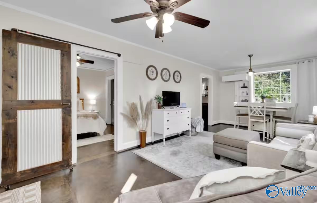 Living room with a rustic sliding barn door, light-colored seating, and adjoining dining area by a large window.