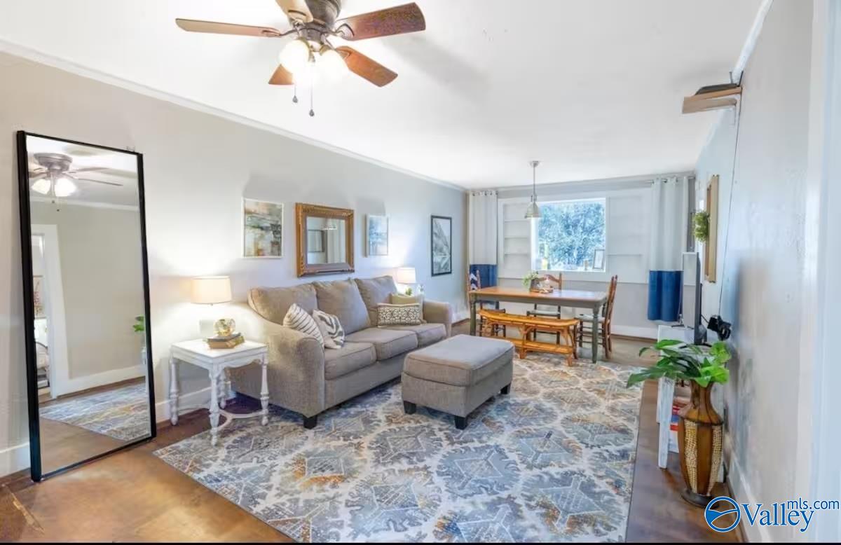Living room with a gray sofa and ottoman, patterned rug, ceiling fan, and dining area by a large window.