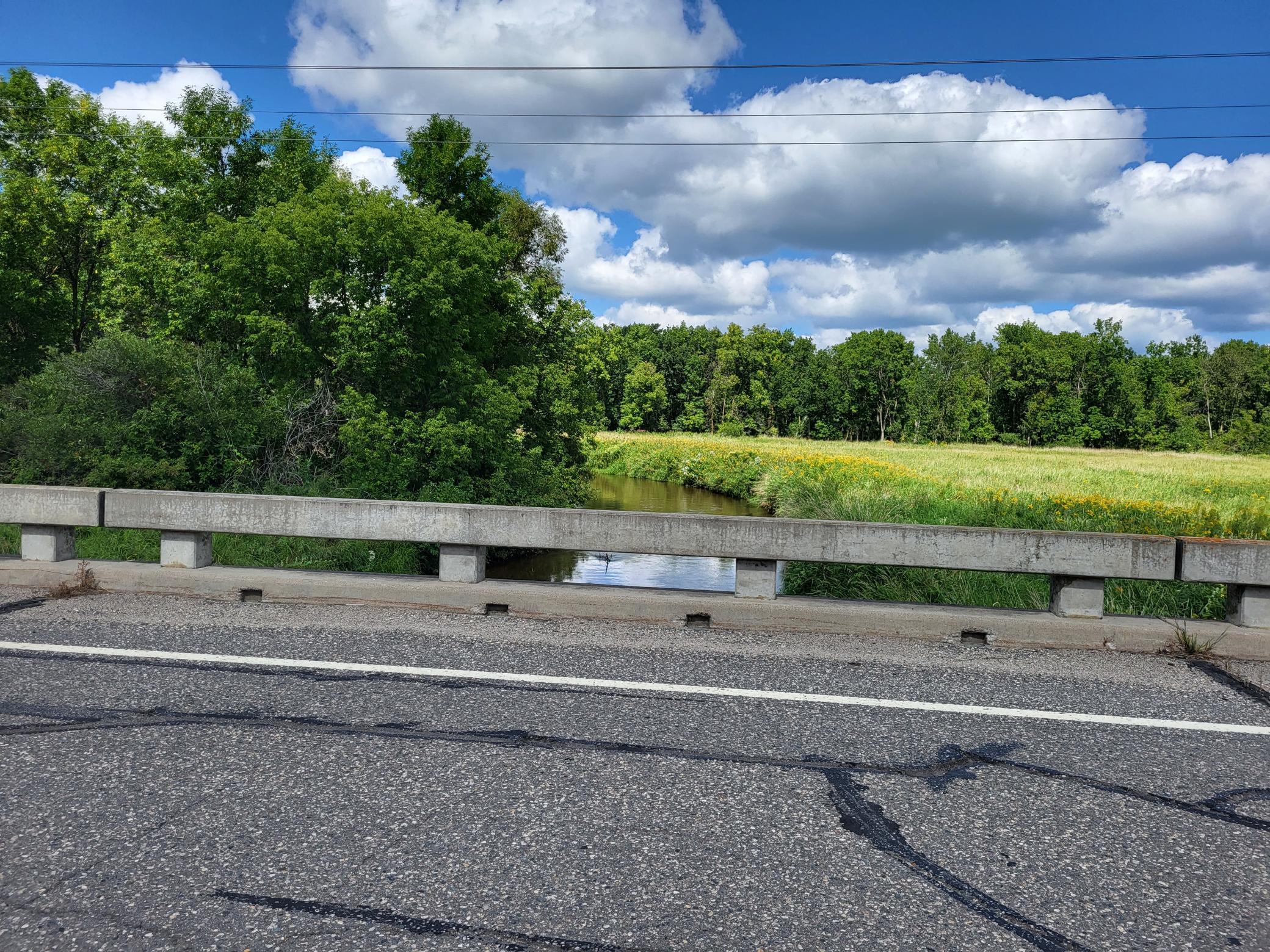 Rural land with a creek between a grassy meadow and dense woods, viewed from a paved bridge.