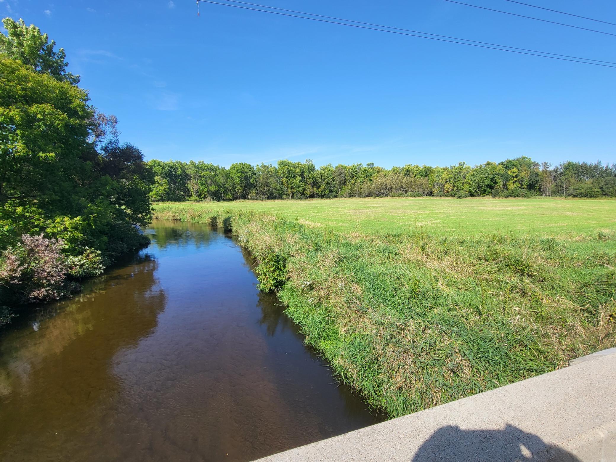 Open field beside a winding creek, viewed from a bridge with trees along the horizon.
