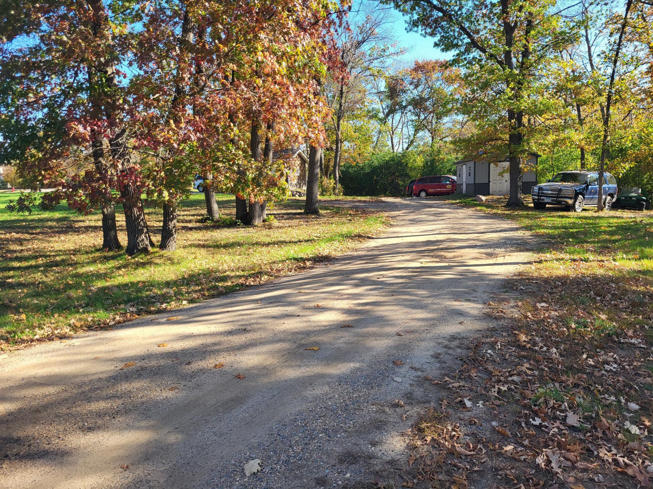 Gravel driveway winding through an autumn lot, with mature trees, a shed, and parked vehicles.