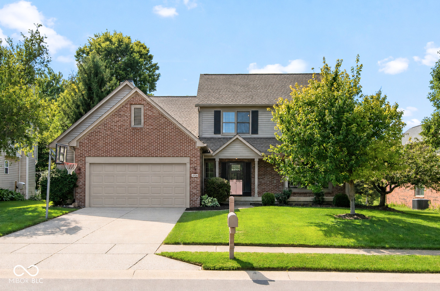 Two-story brick house with an attached garage, mature trees, and a basketball hoop by the driveway.