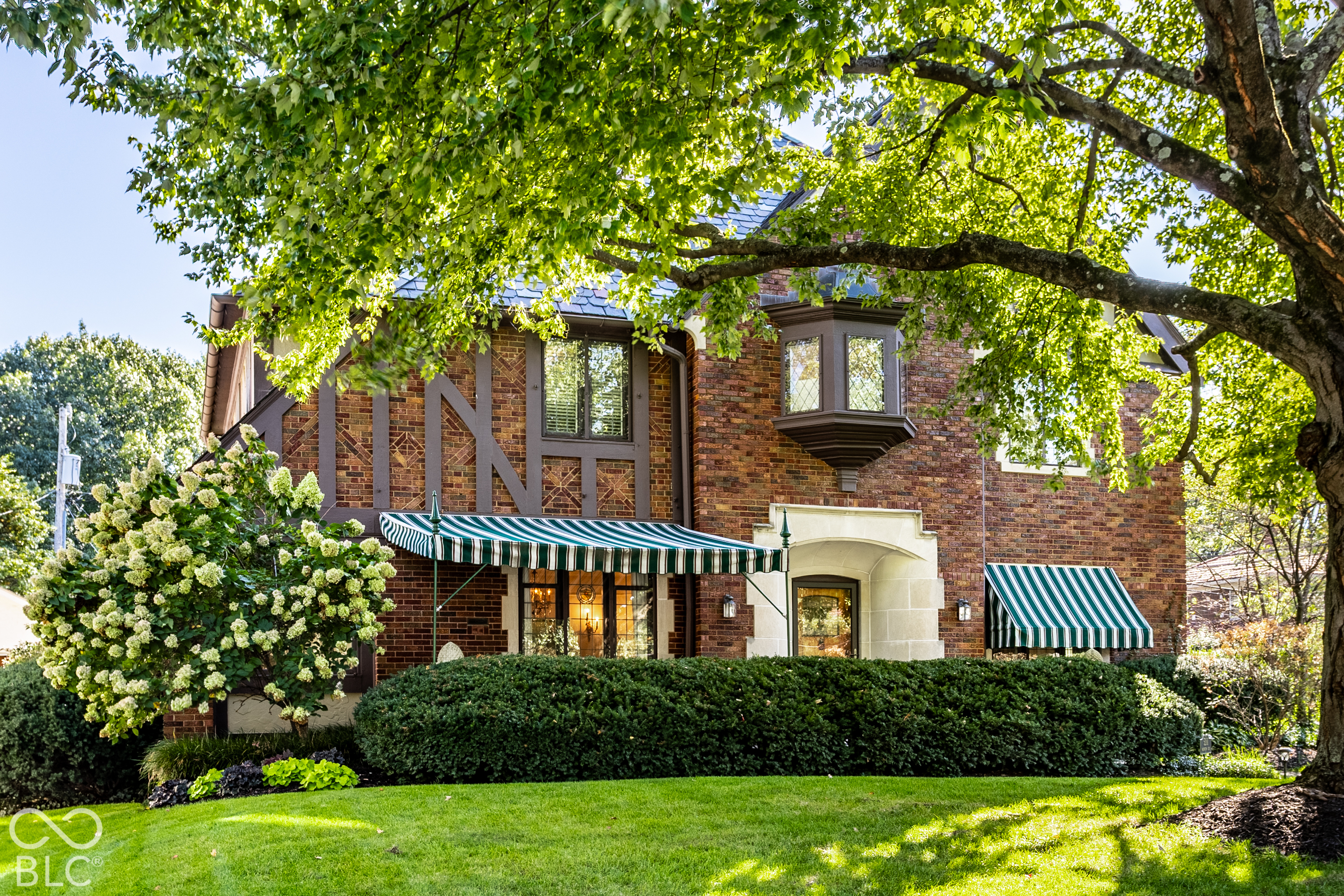 Brick home with striped awnings, a stone entry surround, and mature trees shading the landscaped lawn.