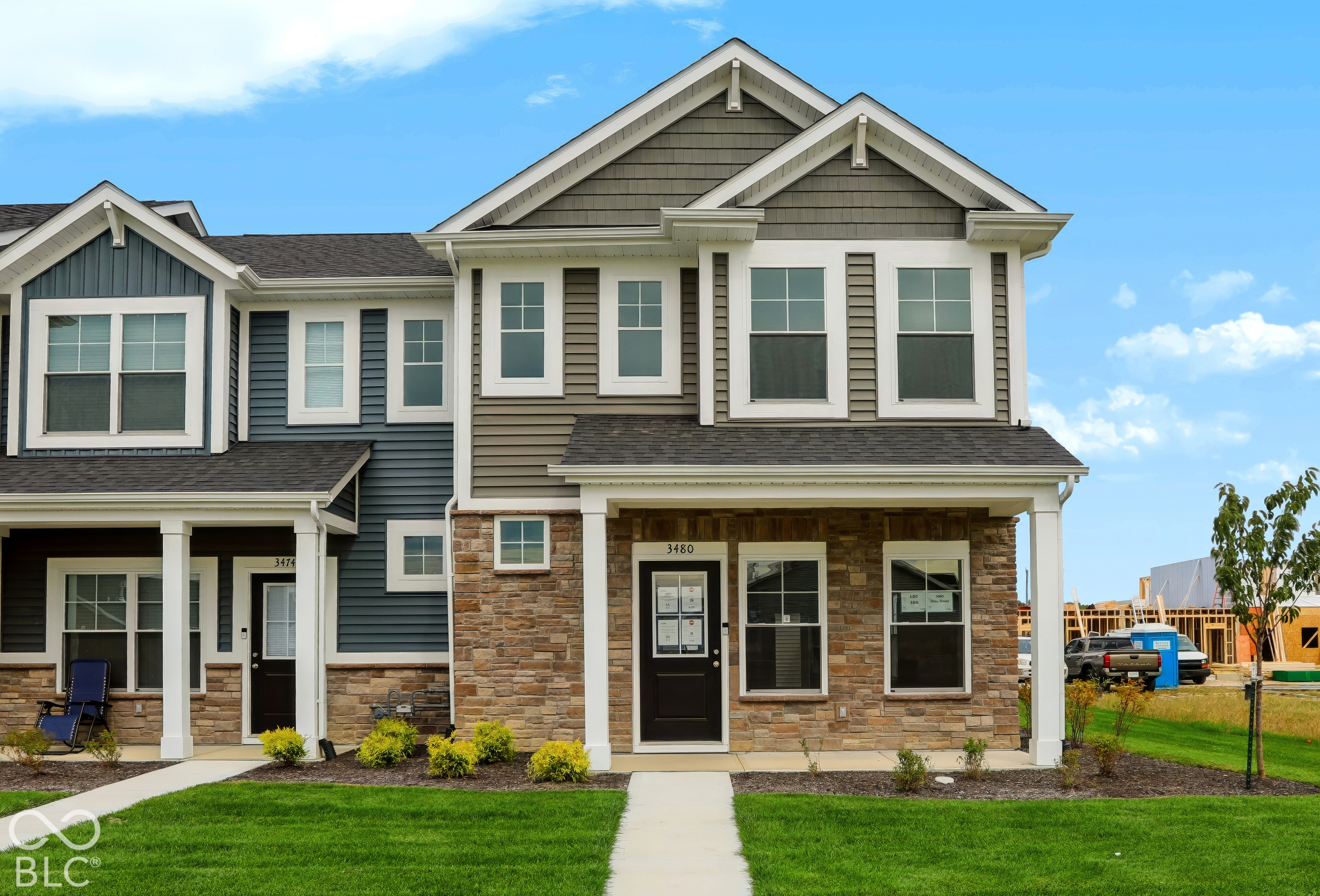 Attached two-story homes with stone-front porches, white trim, and green lawns beside a construction site.