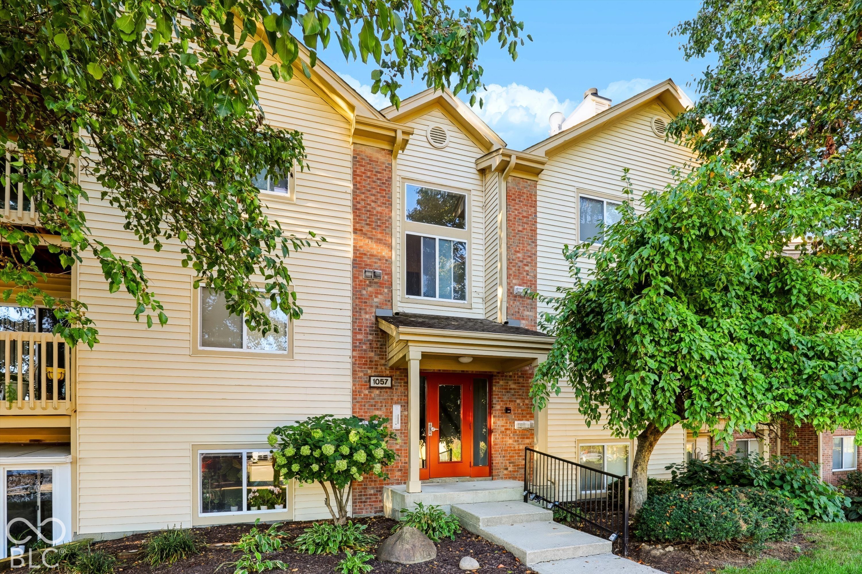 Two-story cream-sided condominium with brick accents, a bright red-orange door, and leafy landscaping.