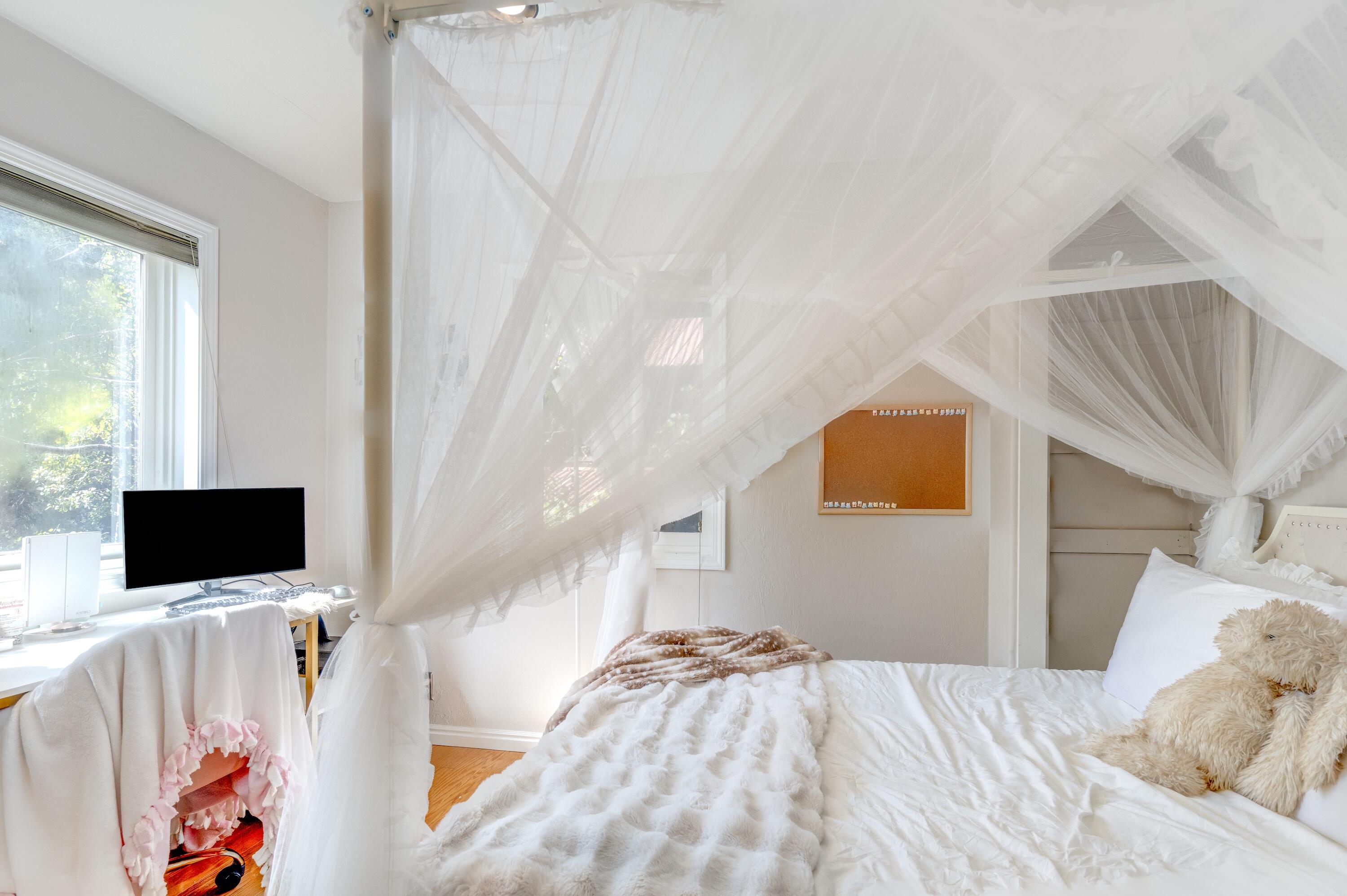 Bright bedroom with a white canopy bed, fluffy bedding, and a desk beside the window.