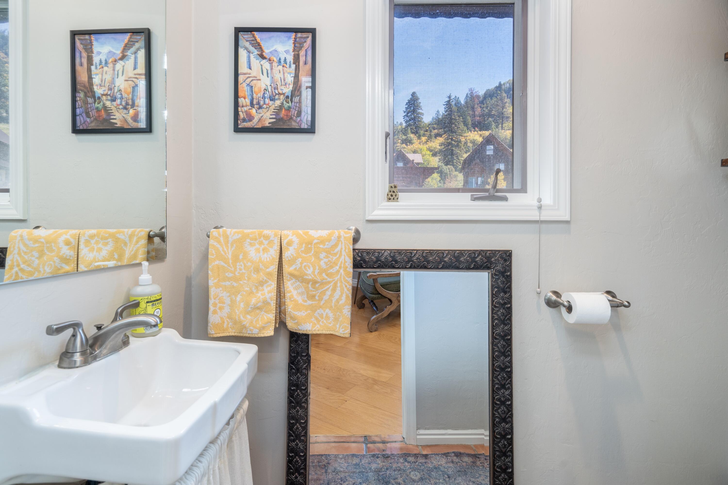 Bathroom vanity with patterned yellow towels, framed artwork, and a window overlooking wooded homes.