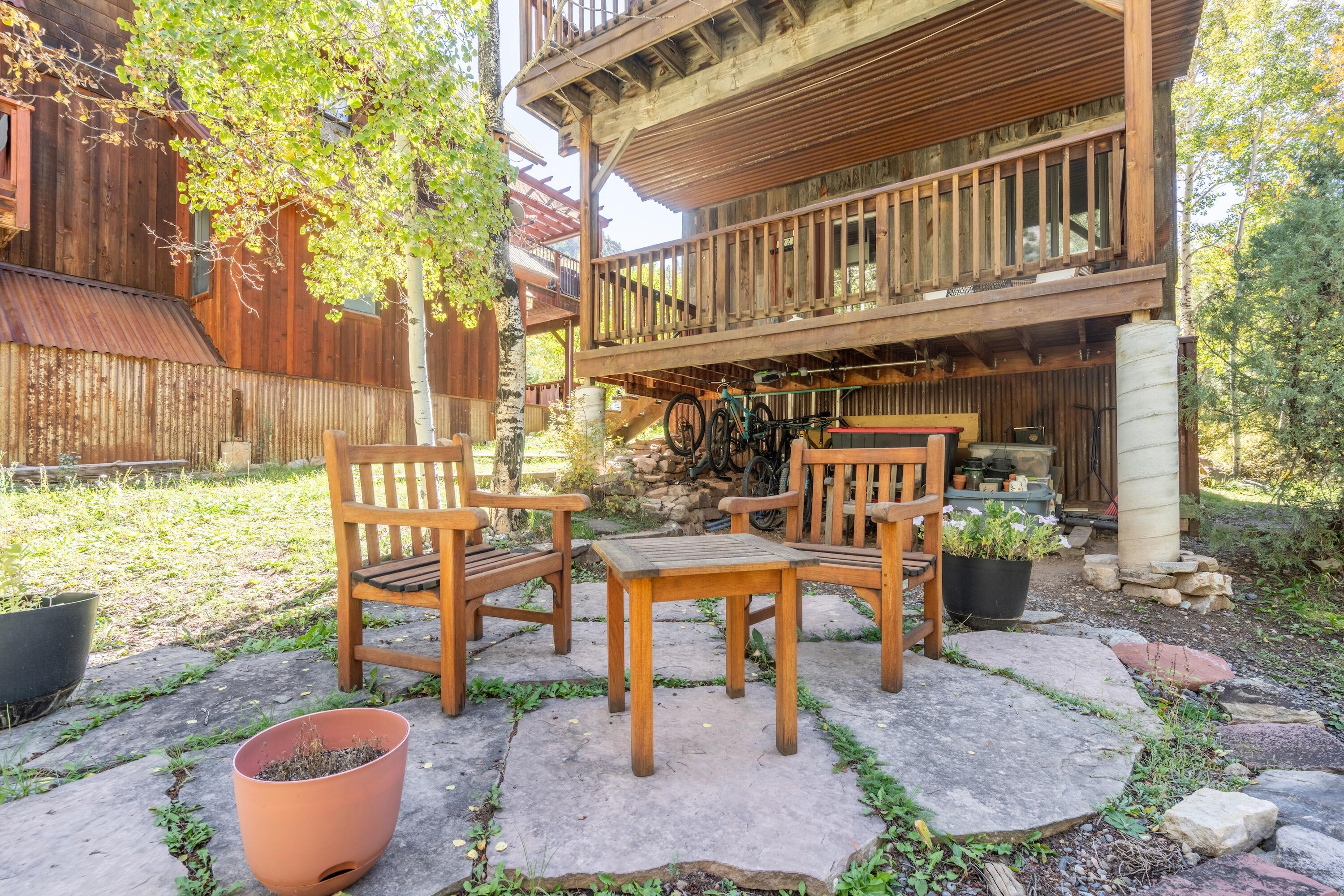 Stone patio with wooden seating beneath a raised deck, surrounded by trees and garden plants.