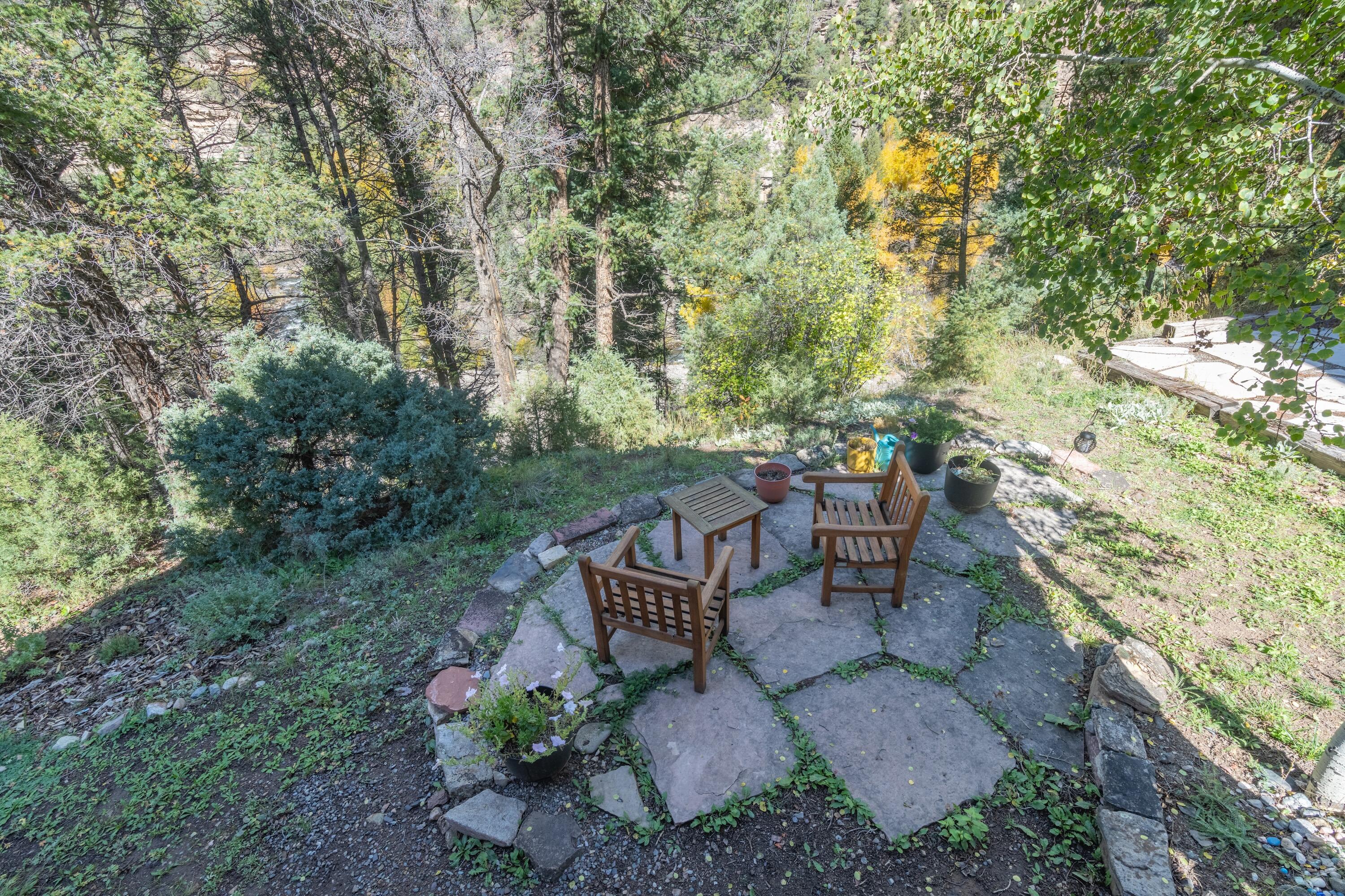 Stone patio with wooden chairs and table surrounded by trees and shrubs.