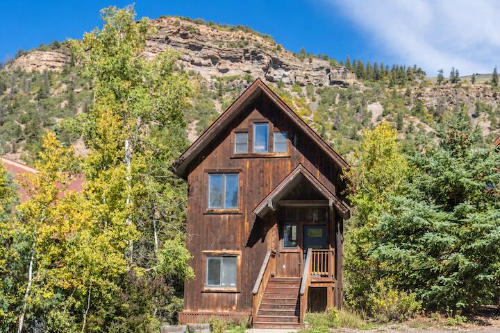 Wood-sided mountain home with a steep gabled roof and front porch beneath a rocky hillside.
