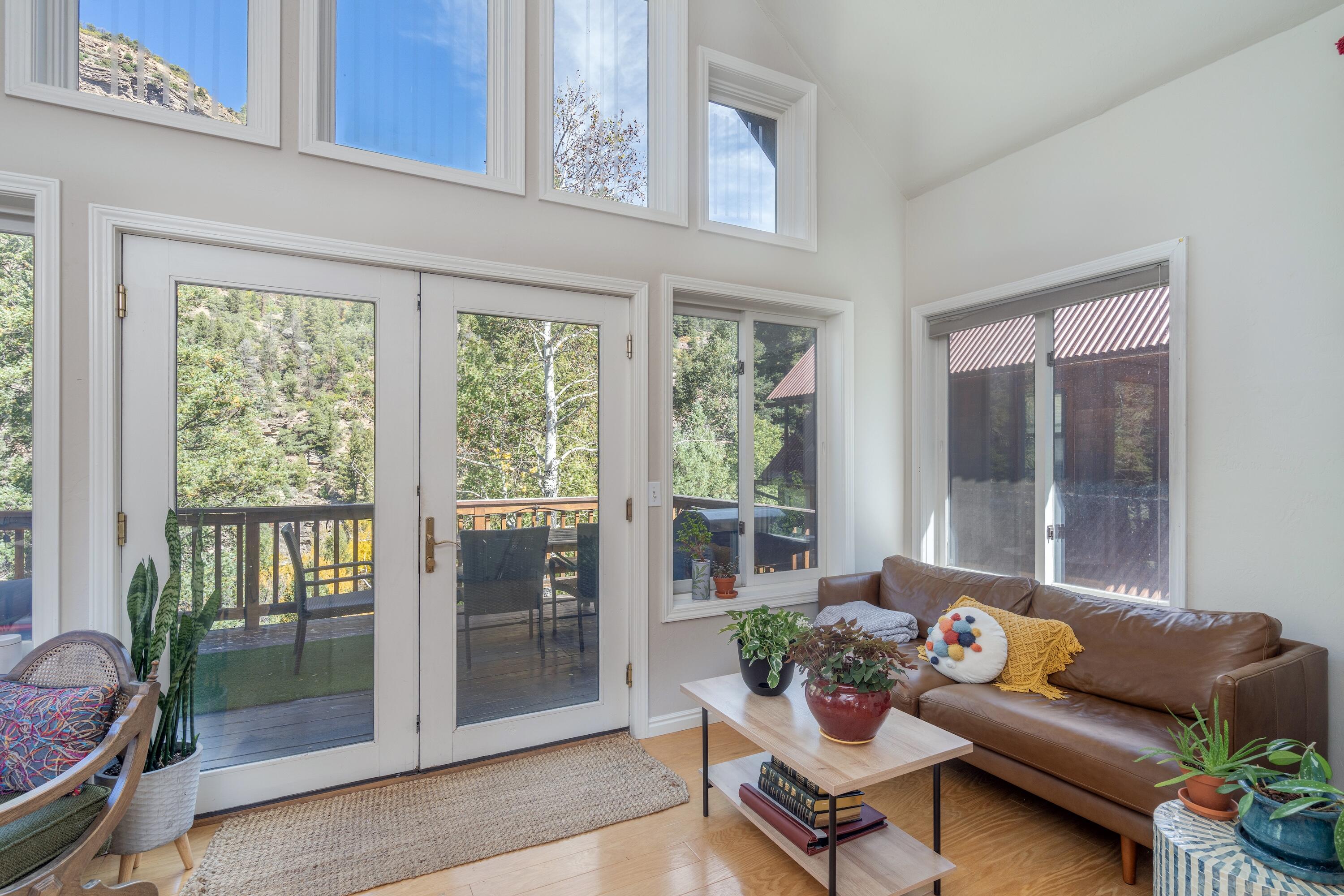 Living room with tall windows, a brown leather sofa, and mountain greenery beyond the deck.