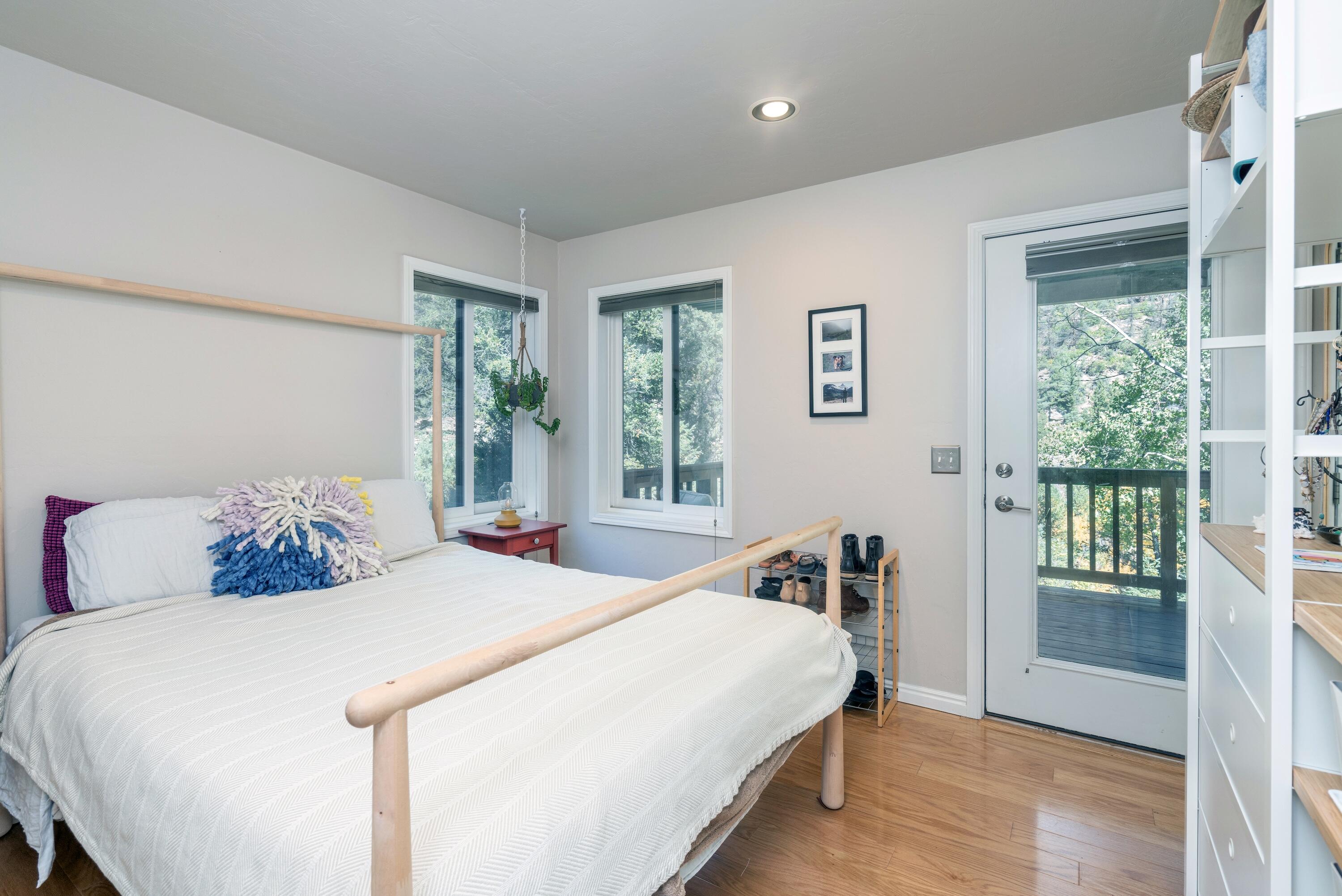 Bedroom with a white bed, colorful pillows, and a glass door opening onto a wooded balcony.
