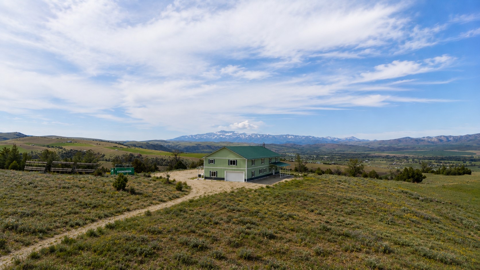 Green two-story building on a rural hillside, with a dirt driveway and snow-capped mountains in the distance.