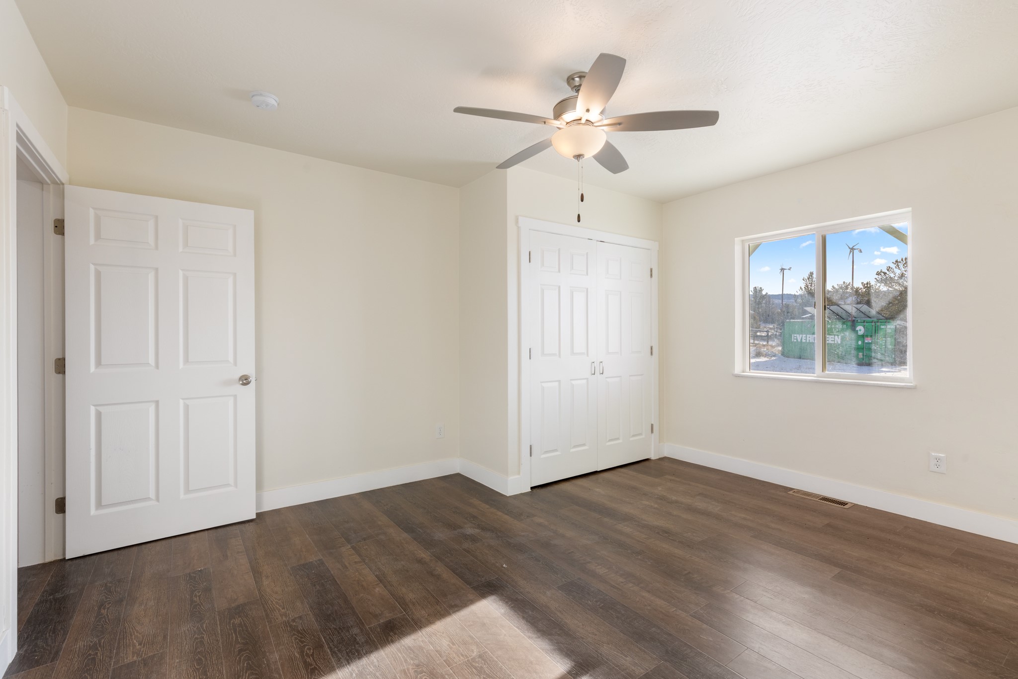 Empty bedroom with dark plank flooring, white closet doors, and a ceiling fan; snow is visible through the window.