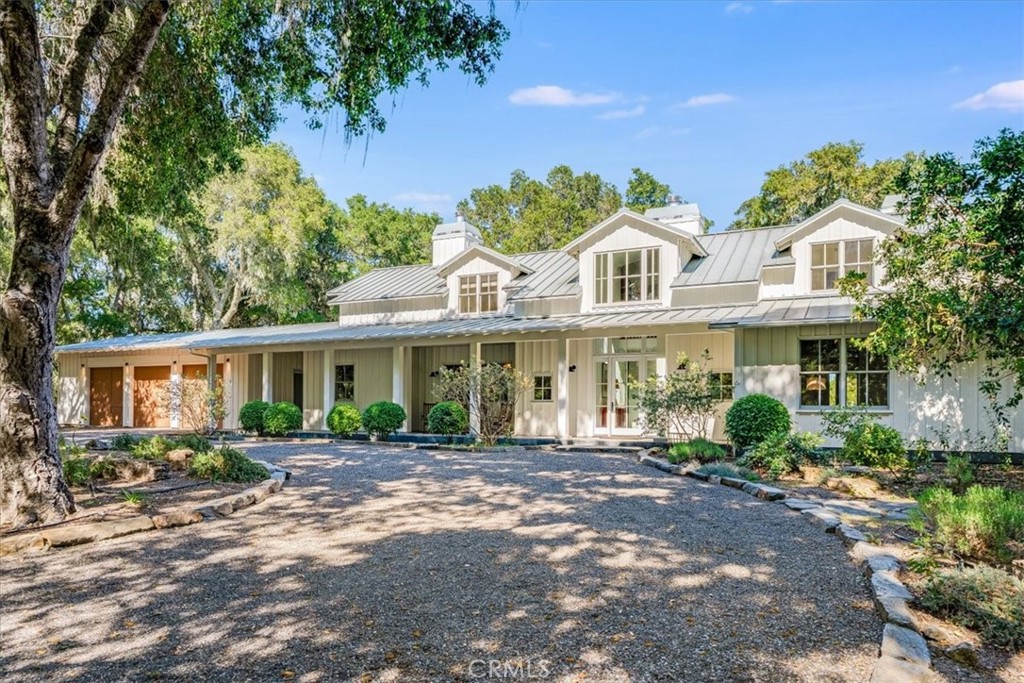 White-and-sage home with a metal roof and dormer windows, viewed from a tree-shaded gravel drive.