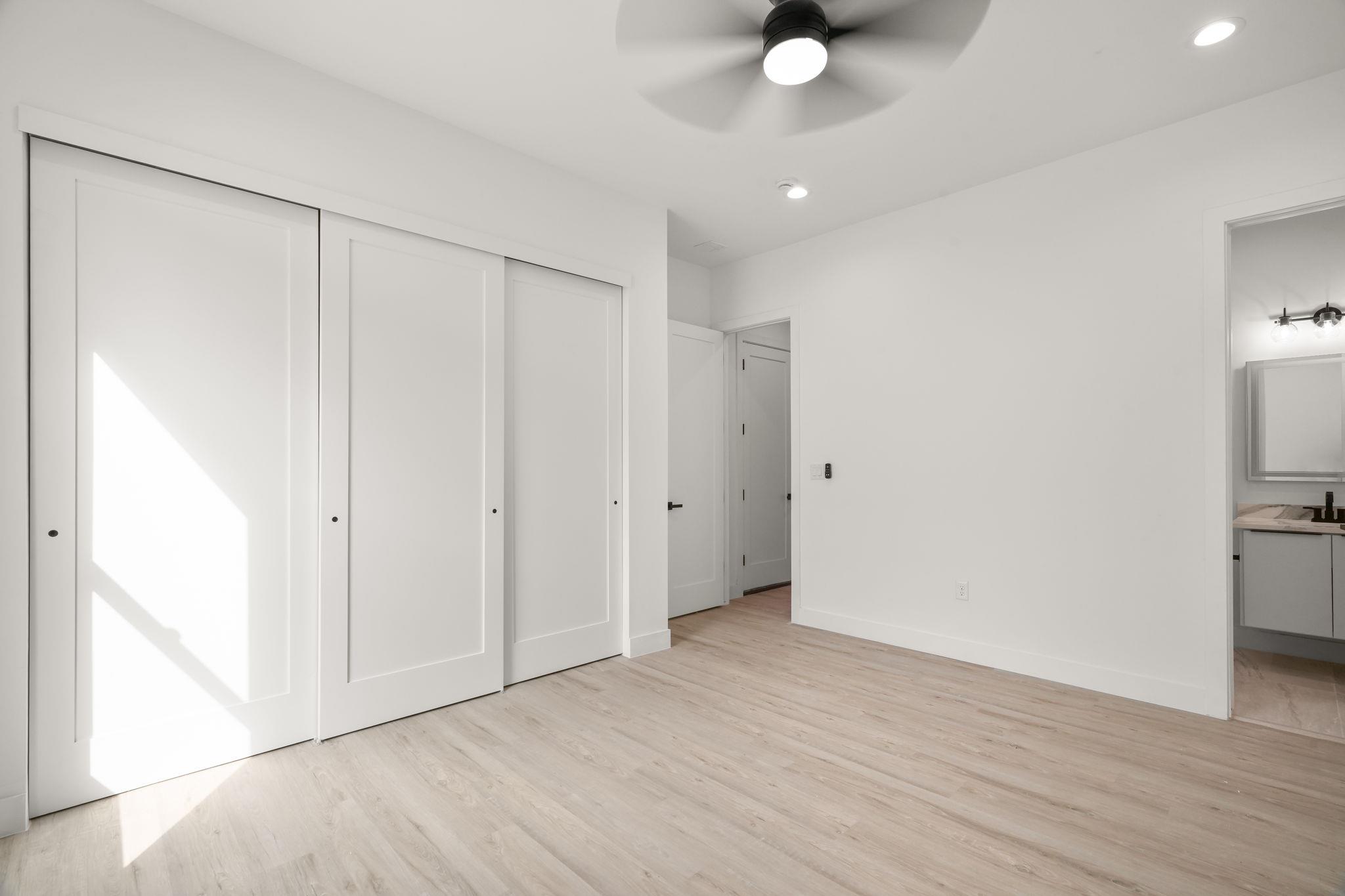 Vacant bedroom with white sliding closet doors, light wood flooring, and a ceiling fan.