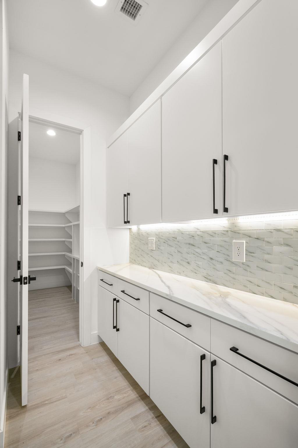 Kitchen with white cabinetry, marble-look counters and backsplash, opening to a shelved pantry.