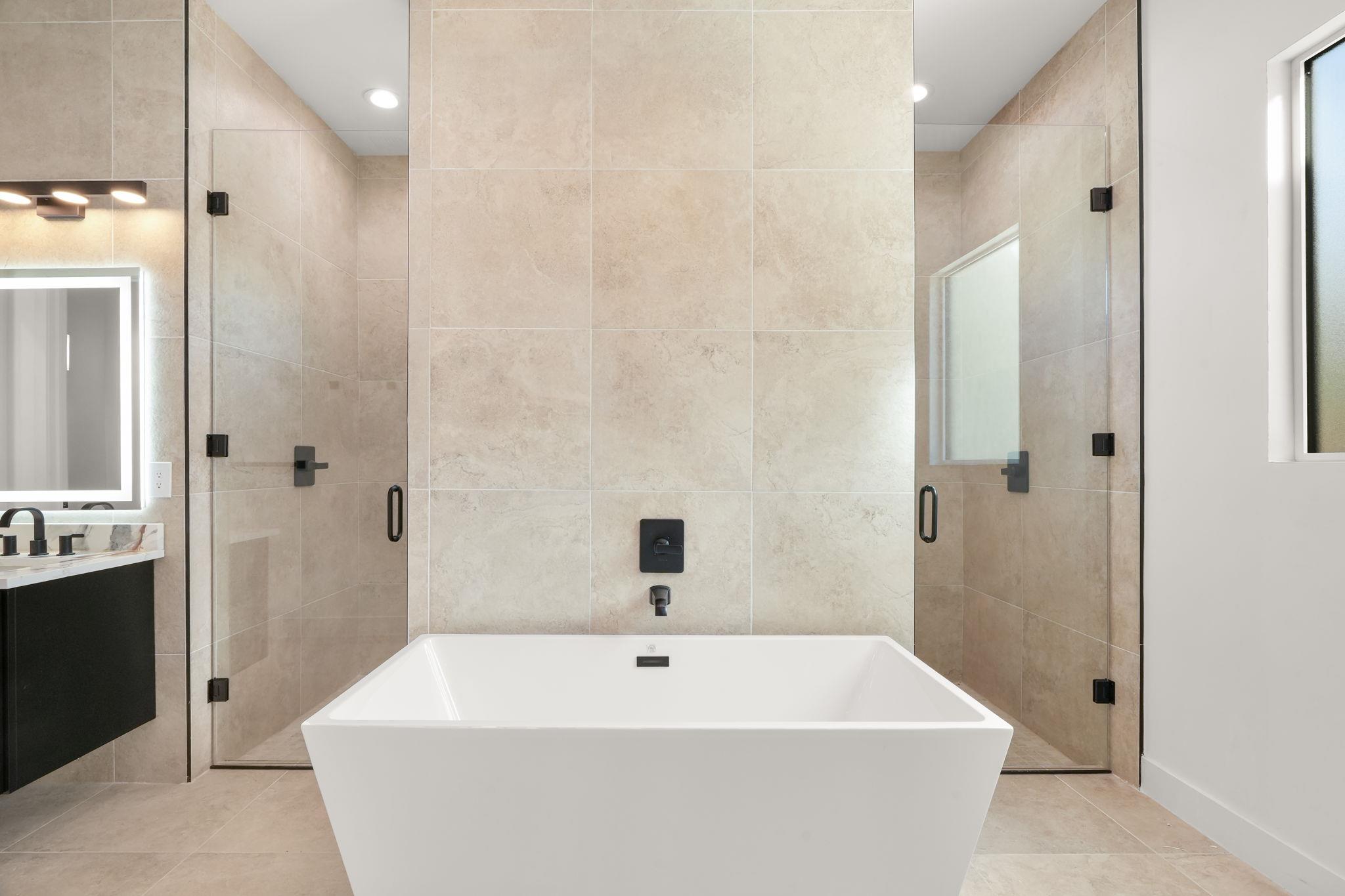 Modern bathroom with a freestanding soaking tub, twin glass showers, beige tile, and a backlit mirror.