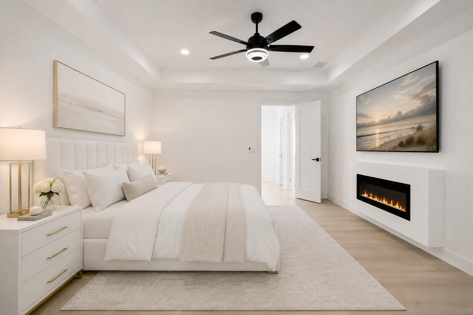 Staged white bedroom with an upholstered bed, black ceiling fan, and modern linear fireplace.