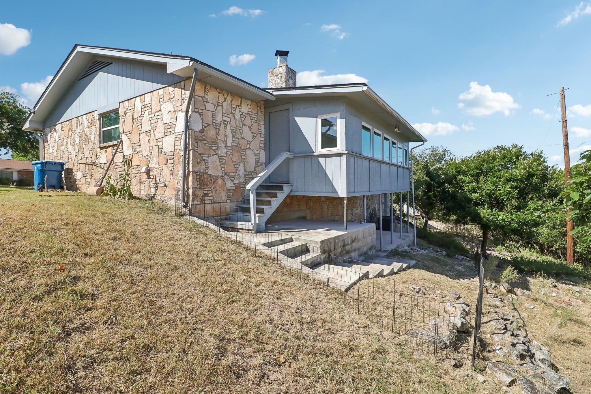Stone-and-gray-sided home with exterior stairs and a covered patio on a sloping yard.