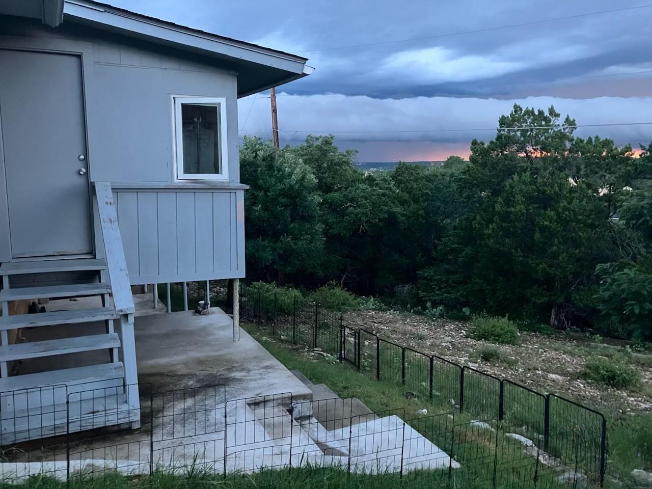 Raised light-colored home with porch and fenced patio overlooking trees beneath a dramatic dusk sky.