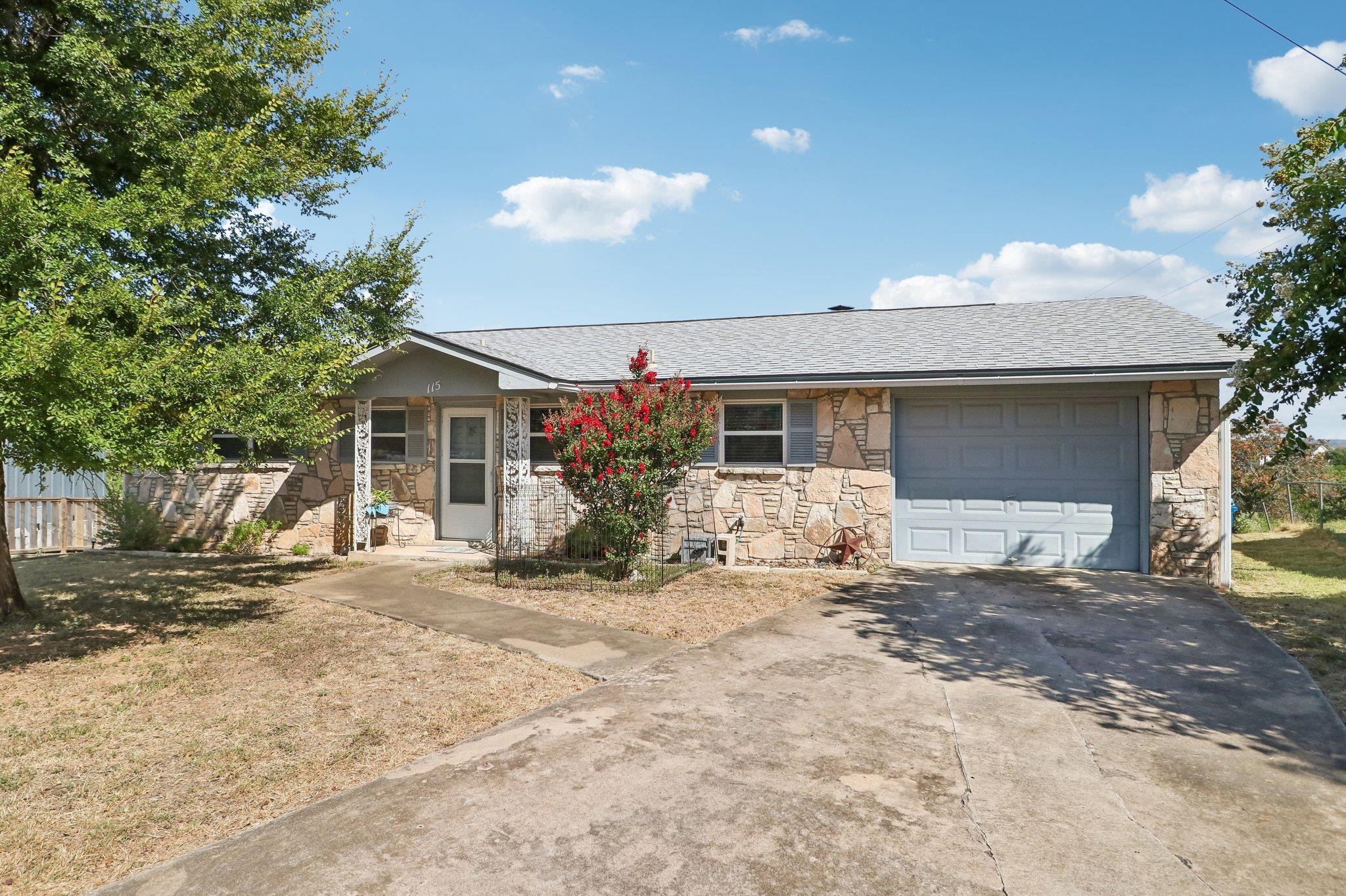 Single-story stone-front house with an attached garage and red flowering shrub beside the entrance.