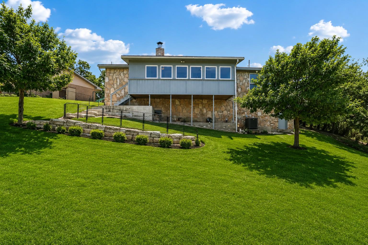 Raised stone-and-siding home above a fenced patio and broad green lawn.
