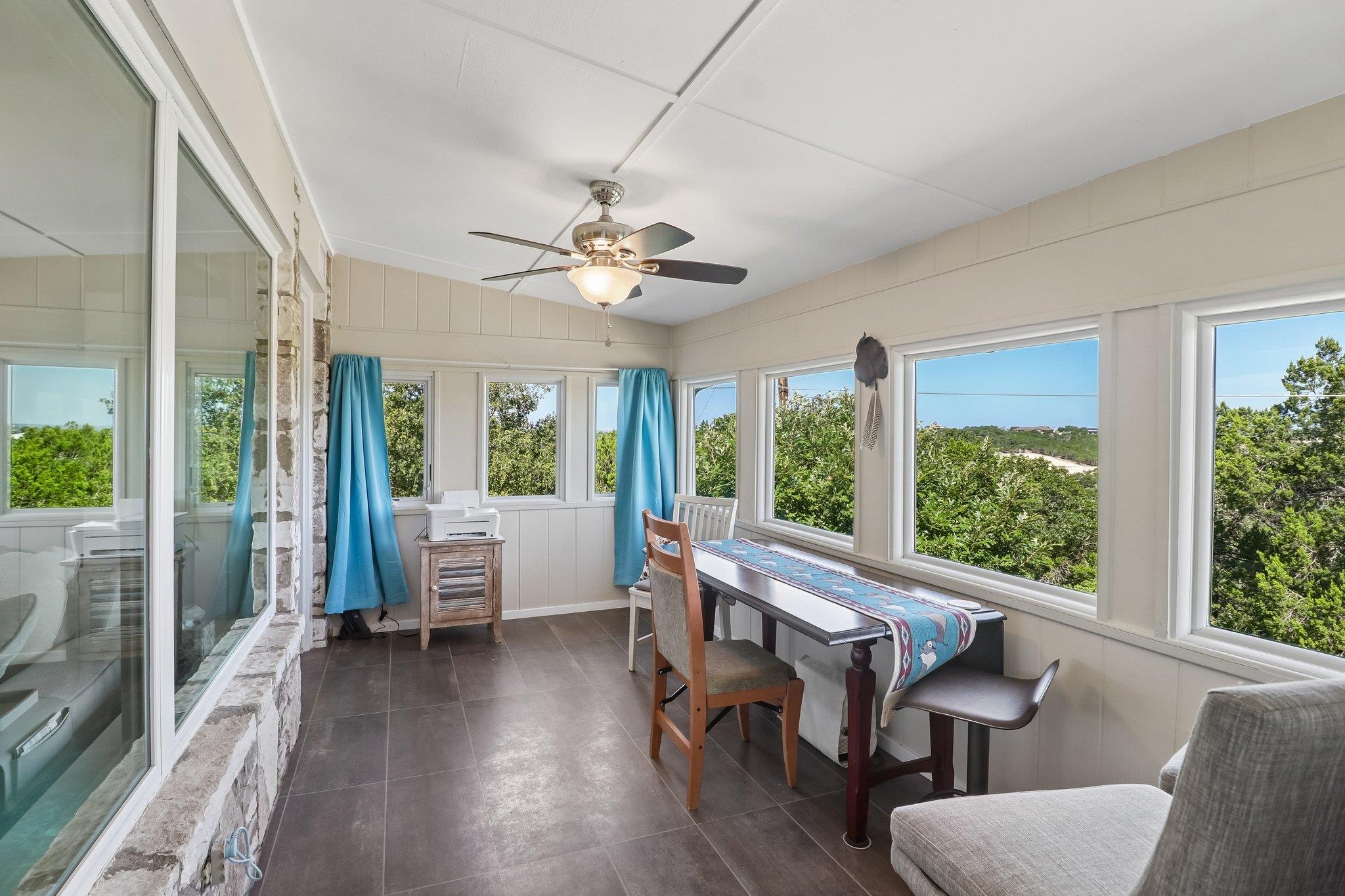 Furnished sunroom with a long table, blue curtains, and large windows overlooking greenery.