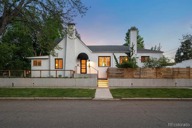 White gabled house with lit windows, tall chimneys, and a horizontal wood fence at dusk.